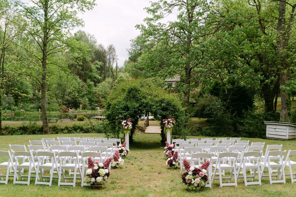 Outdoor wedding setup with white chairs and flower arrangements on a grassy lawn, surrounded by trees and a pond in the background.