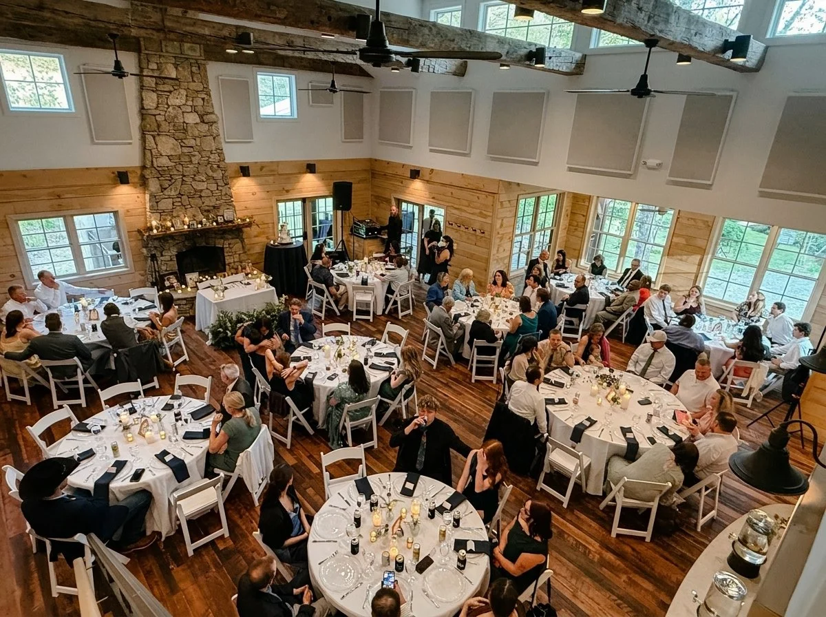 A large banquet hall decorated for an event with multiple round tables set with black napkins, candles, and glasses, filled with guests seated and conversing. The room features wooden walls, a stone fireplace, large windows, and ceiling fans.