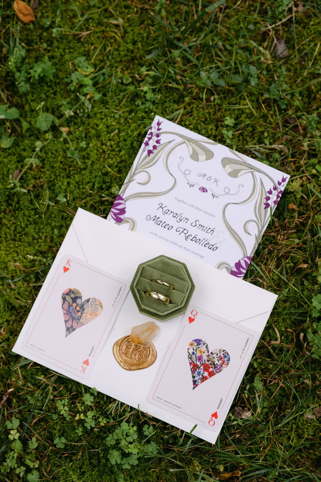 A wedding invitation card with purple floral design, two playing cards (King of hearts and Queen of hearts) with decorative heart-shaped artwork, a small green velvet ring box with two gold wedding bands, and a wax seal with a small decorative gem, a