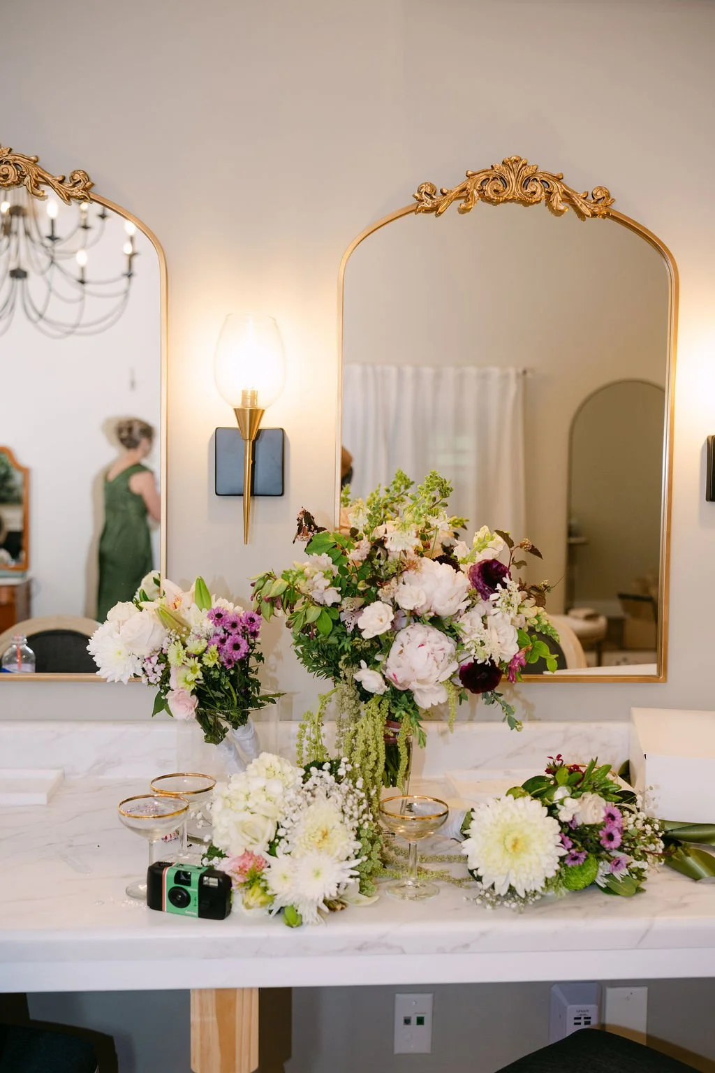 A marble bathroom counter with three floral arrangements in vases, a vintage camera, and glasses, reflected in ornate gold-framed mirrors, with a wall sconce light on, and a person in a green dress in the background.
