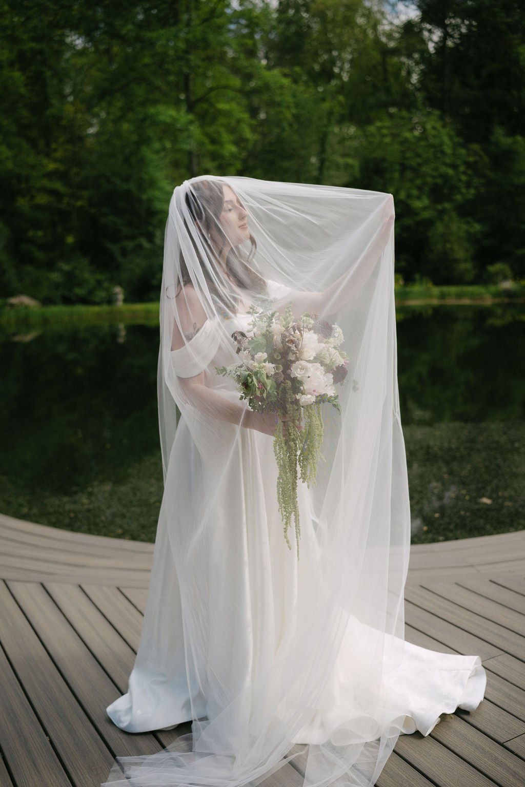 A woman in a wedding dress holds a bouquet of flowers behind a veil outdoors on a wooden deck by a pond, surrounded by green trees.