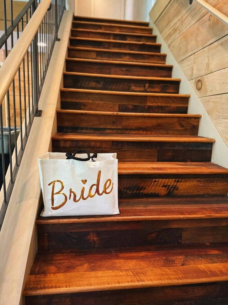 A white tote bag with the word 'Bride' written in gold glitter on it, sitting on a dark wooden staircase with light wood railing.