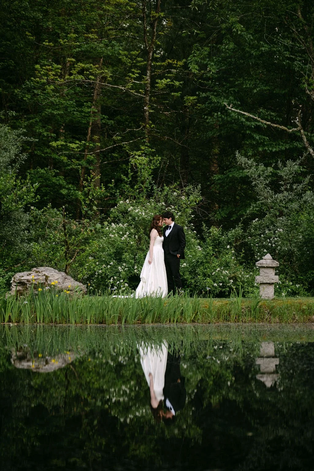 A bride and groom in wedding attire standing close together in a lush green garden, reflected in a pond water in front of them.