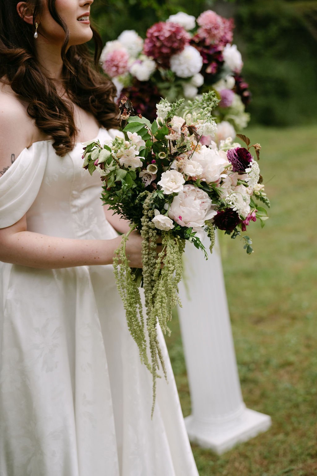 A woman in a white wedding dress holding a bouquet of white, purple, and green flowers outdoors with a floral arch in the background.