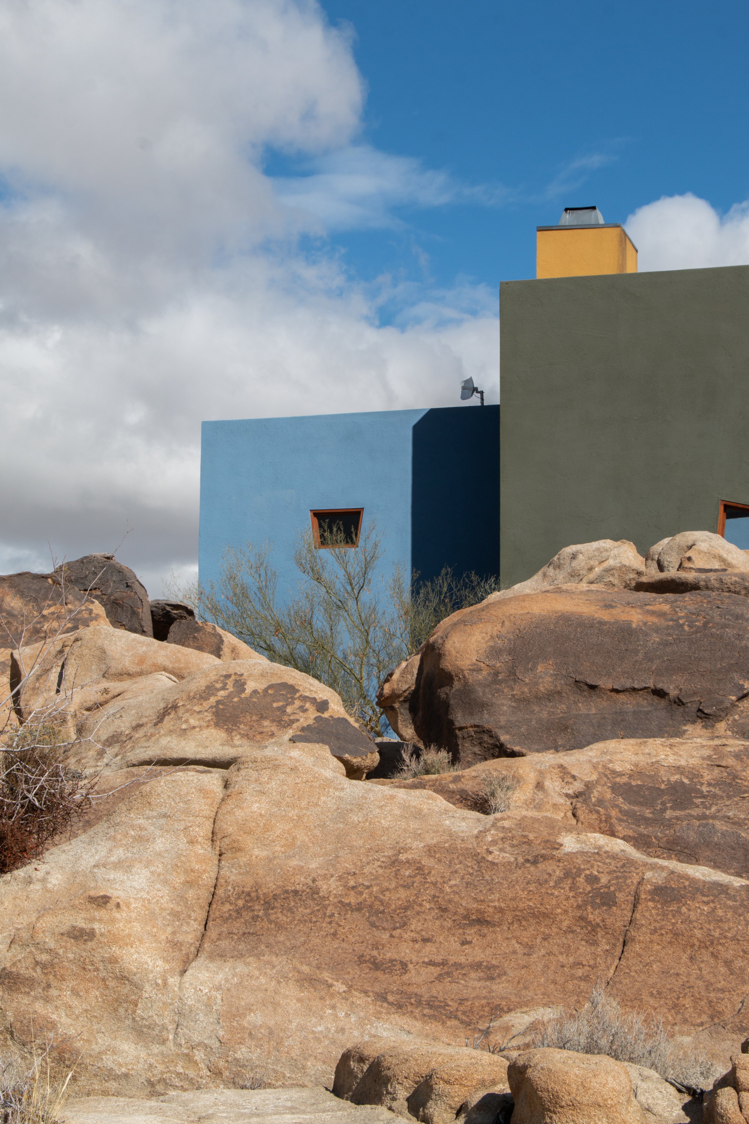 Front view of a modern building with a wooden door, a window with blinds, and a round white exterior light fixture. The building has beige stucco walls and wooden framing, with a small outdoor garden area in the foreground.