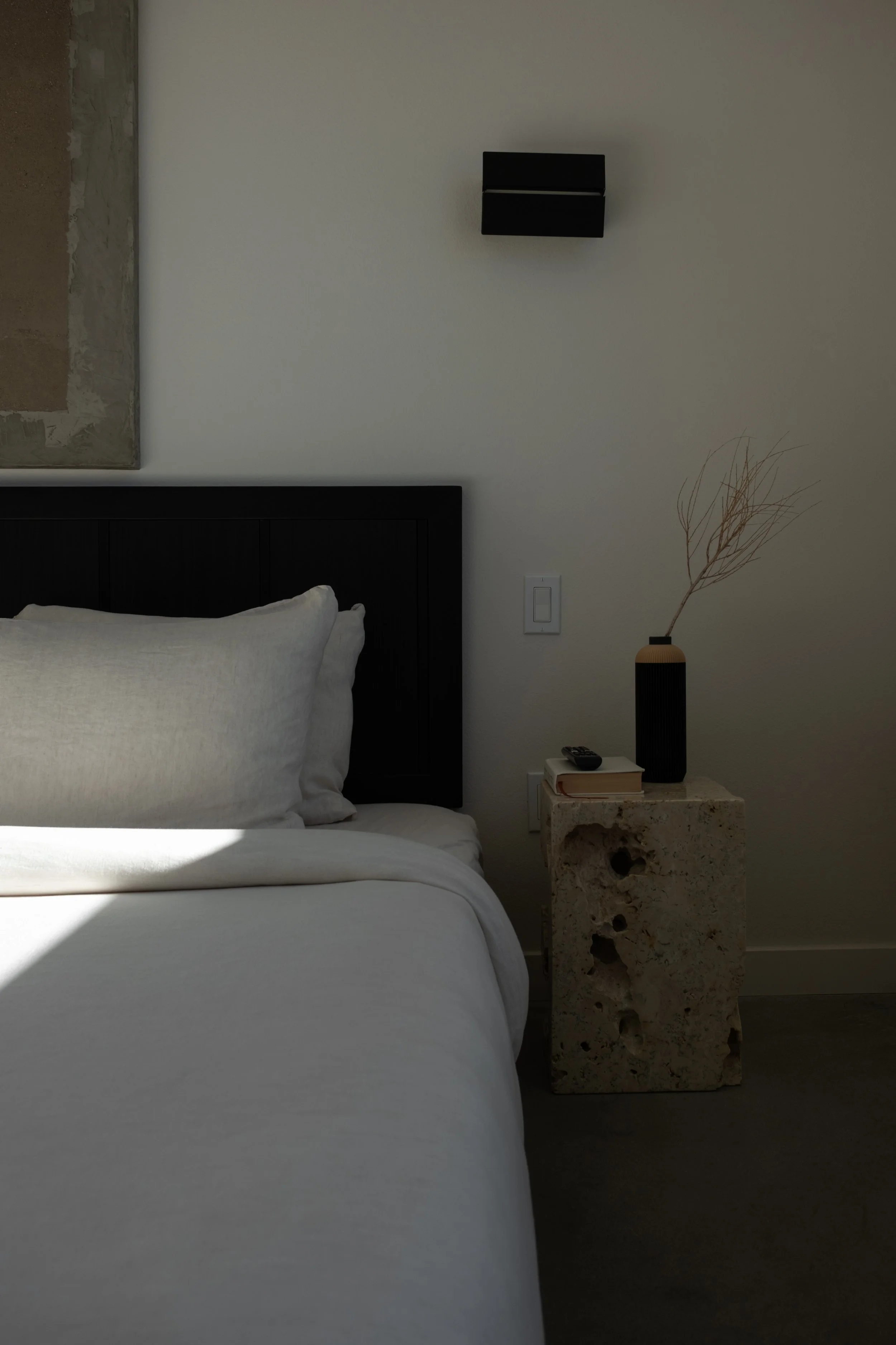 A minimalist bedroom corner with a bed, white bedding, a black headboard, a textured stone nightstand with a black vase and dried branch, a book, and a remote, with a wall-mounted black light fixture and a light switch.