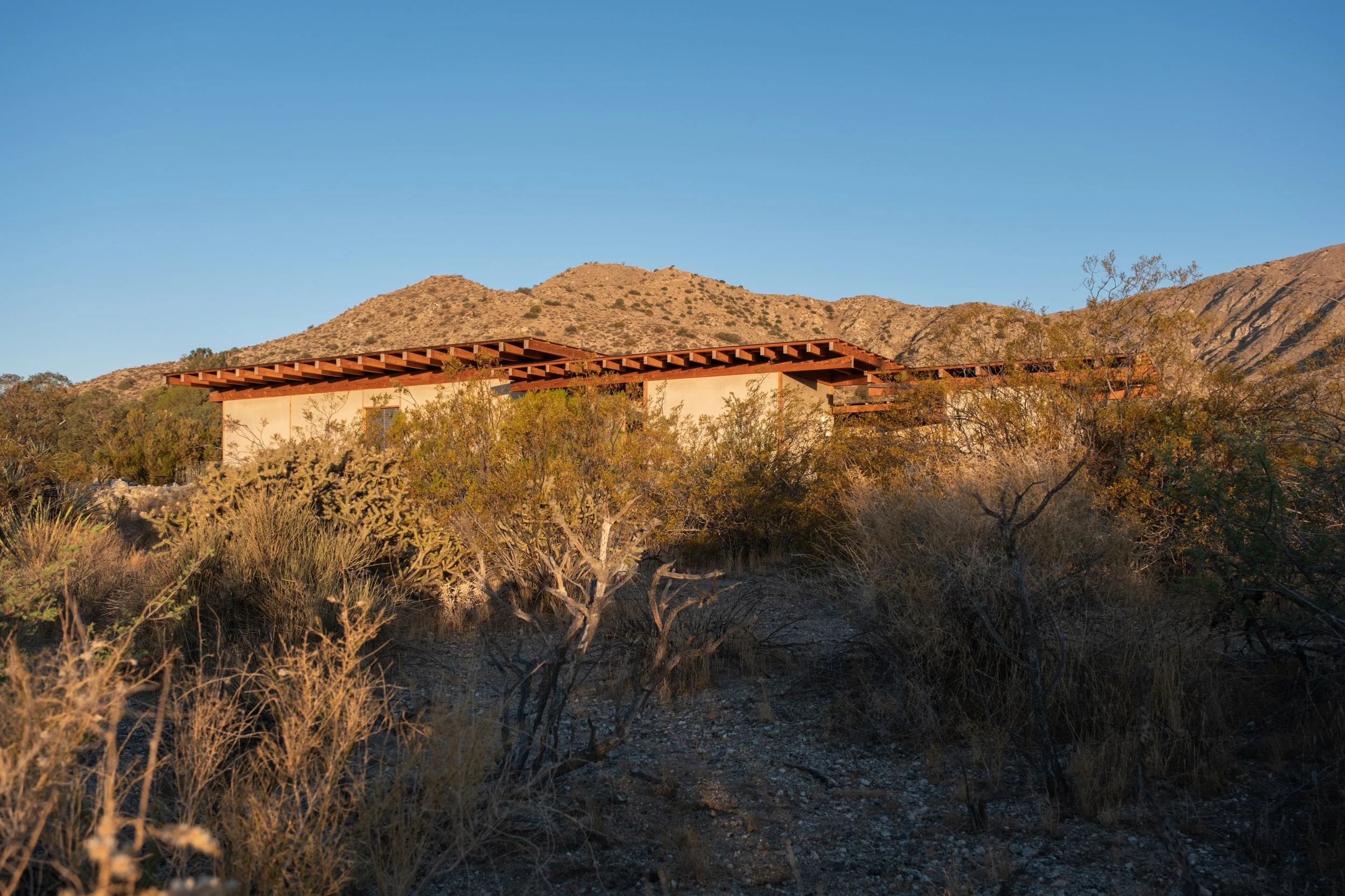 A modern house with a flat roof under construction, located in a desert landscape with dry bushes and mountains in the background, during sunset.