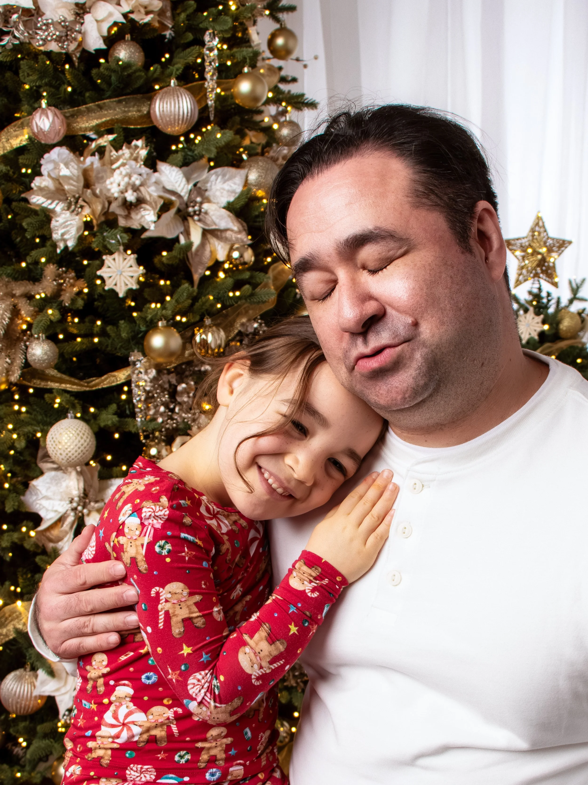 A young girl in Christmas pajamas hugging a man in front of a decorated Christmas tree.