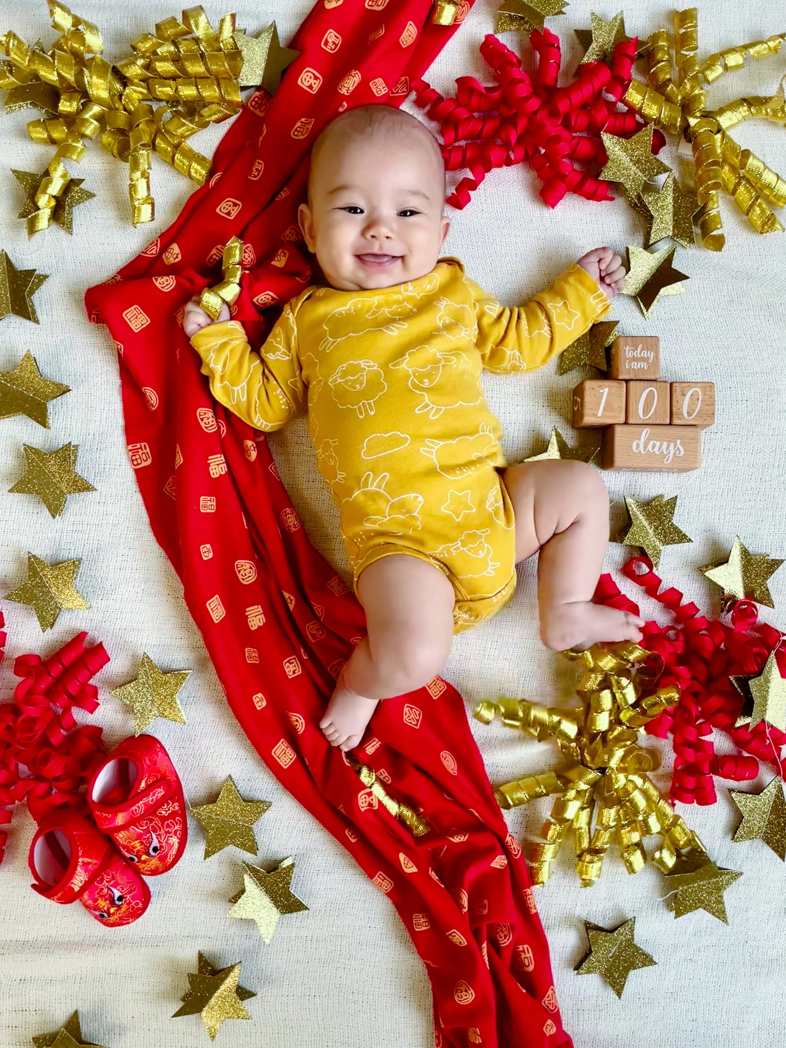 Baby lying on a white surface, surrounded by gold and red decorations, wearing a yellow outfit with cloud and sheep patterns, with a long red cloth, gold and red ribbons, gold stars, red shoes, and wooden blocks displaying "Today I am 100 days."