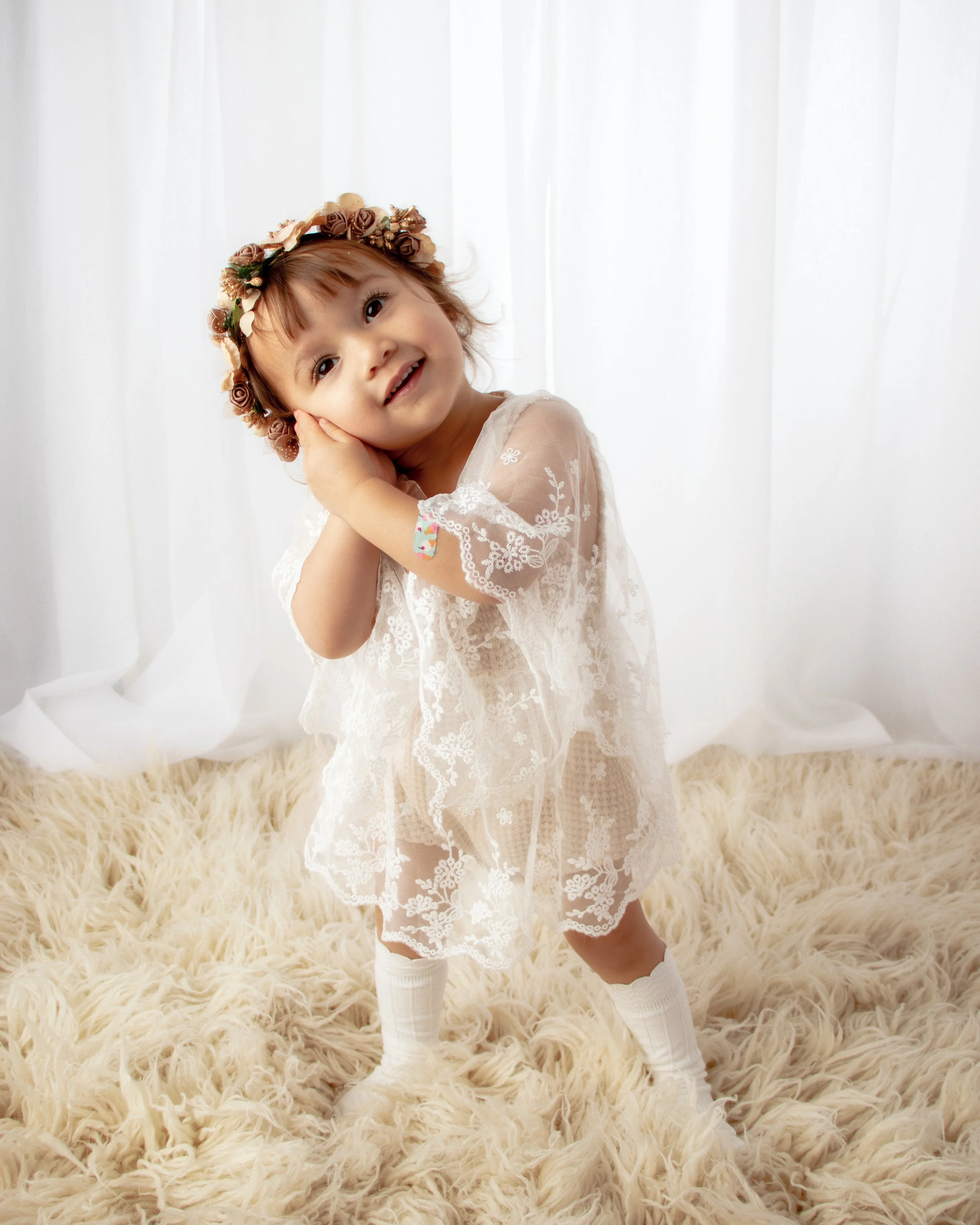 A young girl wearing a lace dress and a floral headband, standing on a fluffy rug in front of white curtains, smiling and posing cheerfully.  Milestone session, Redmond, WA.