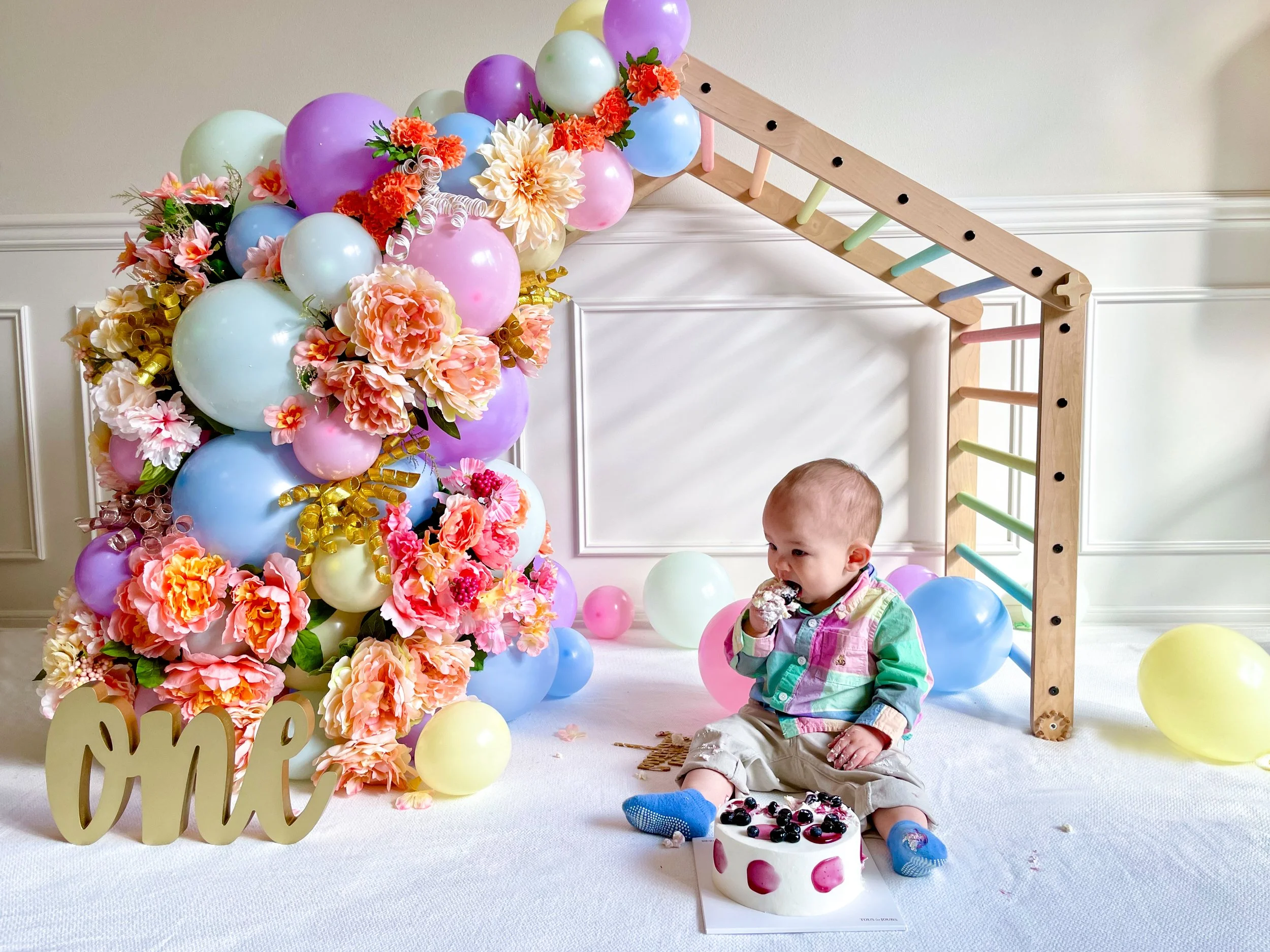 A young child with a colorful shirt eating cake, surrounded by balloons and flowers at a birthday celebration with a wooden arch (Cake Smash Session)