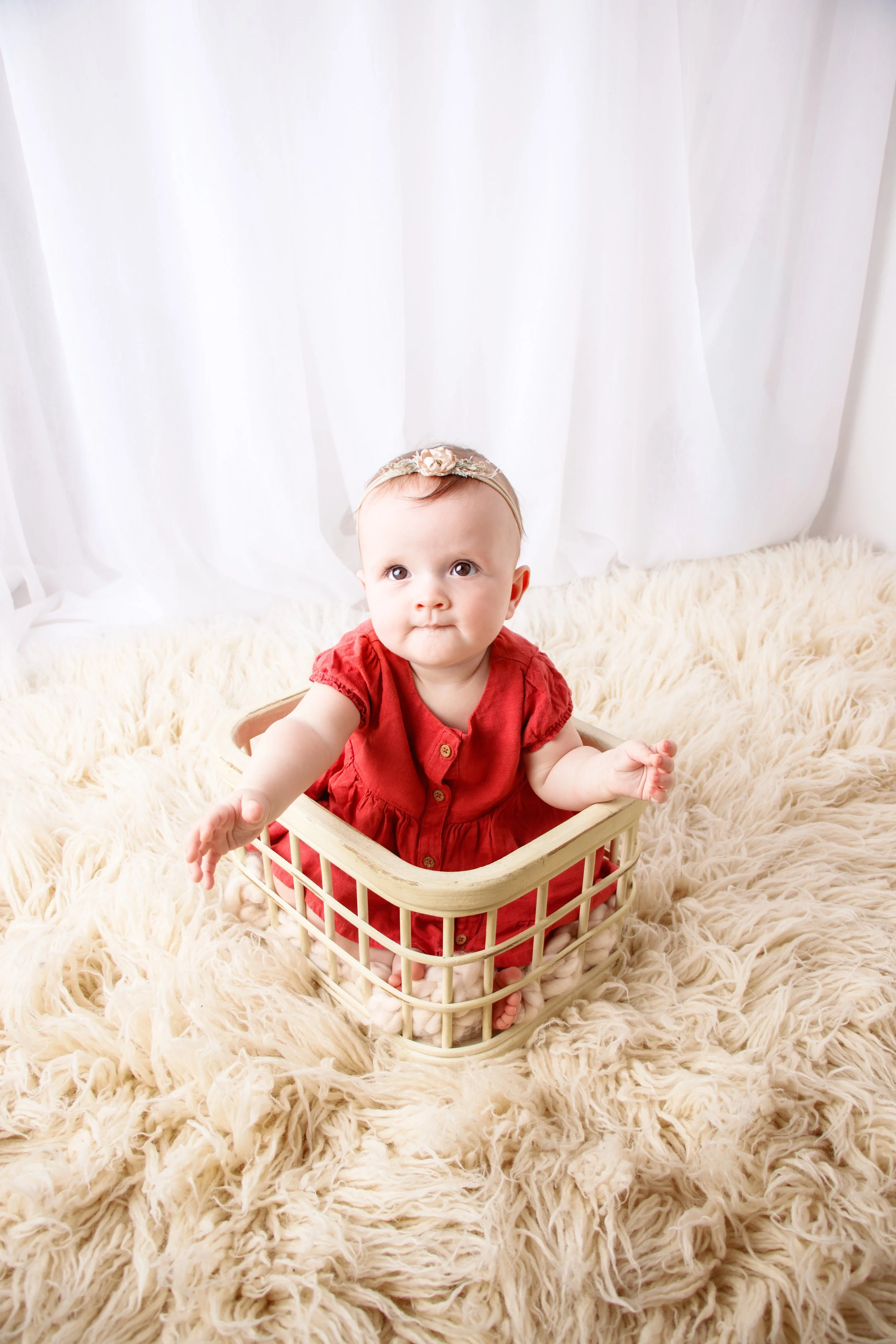 A baby girl sitting in a small basket on a fluffy cream-colored rug, wearing a red dress and a floral headband, with white curtains in the background.  Milestone session, Redmond, WA.