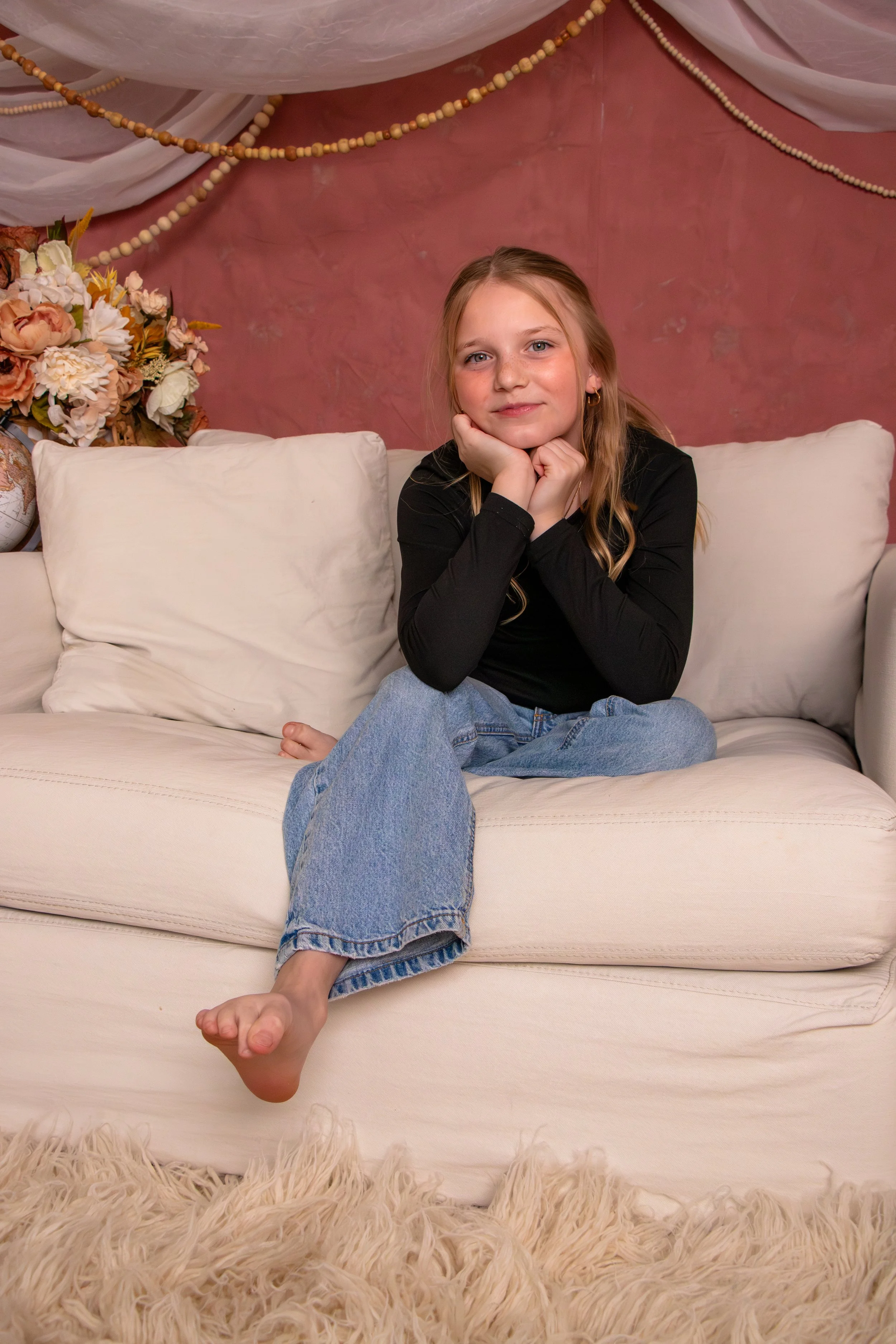 A young girl with blonde hair, wearing a black long-sleeve shirt and jeans, sitting on a beige couch, resting her chin on her hands, looking at the camera. (Family Session)