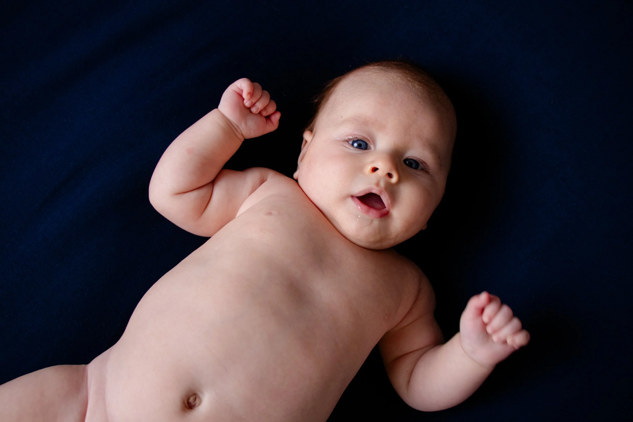 A baby lying on a dark surface with arms raised and mouth slightly open, looking at the camera. (Tummy Session)
