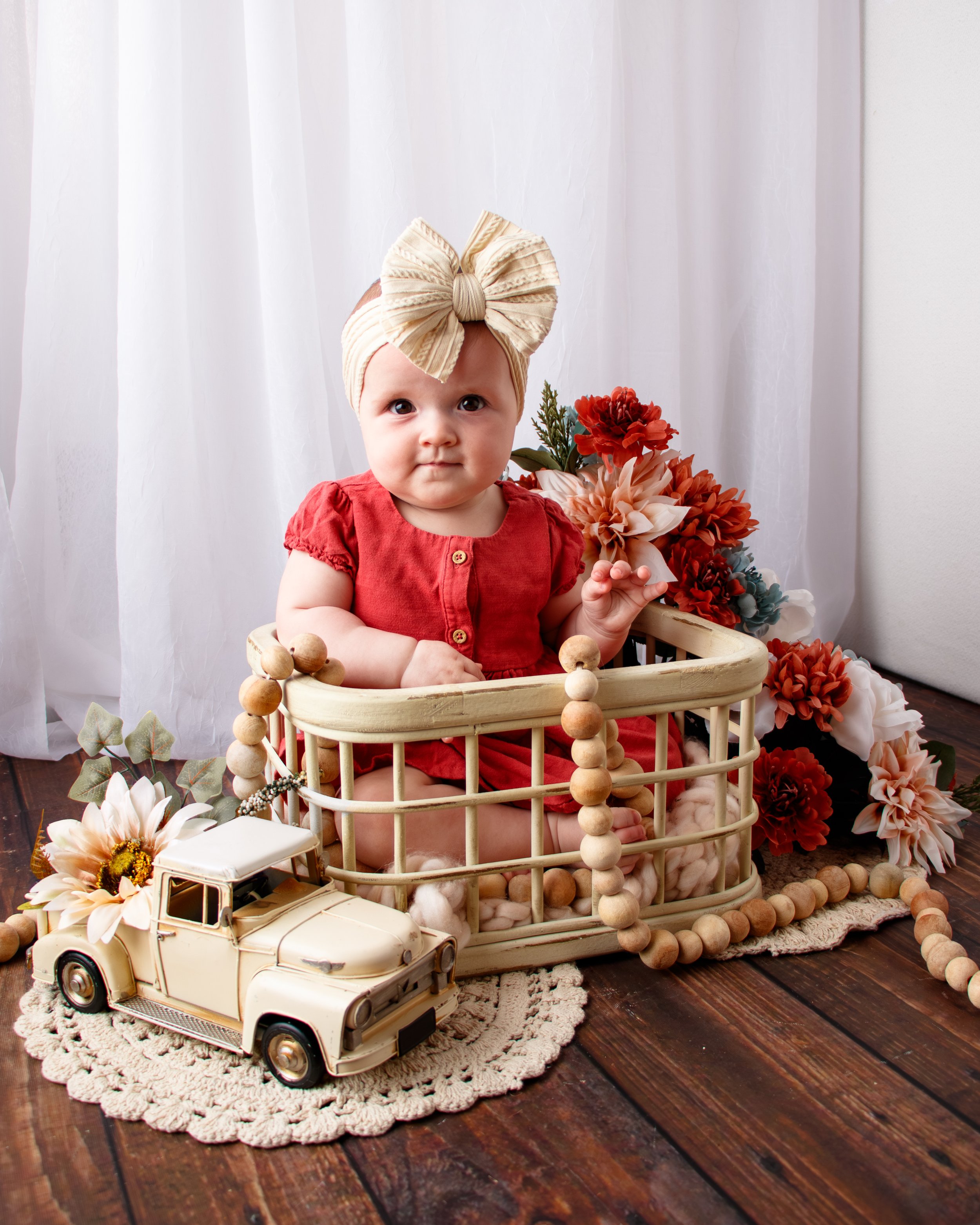 A baby girl sitting in a wooden playpen surrounded by flowers, a toy car, and decorative beads, with white curtains in the background.  Milestone session, Redmond, WA.