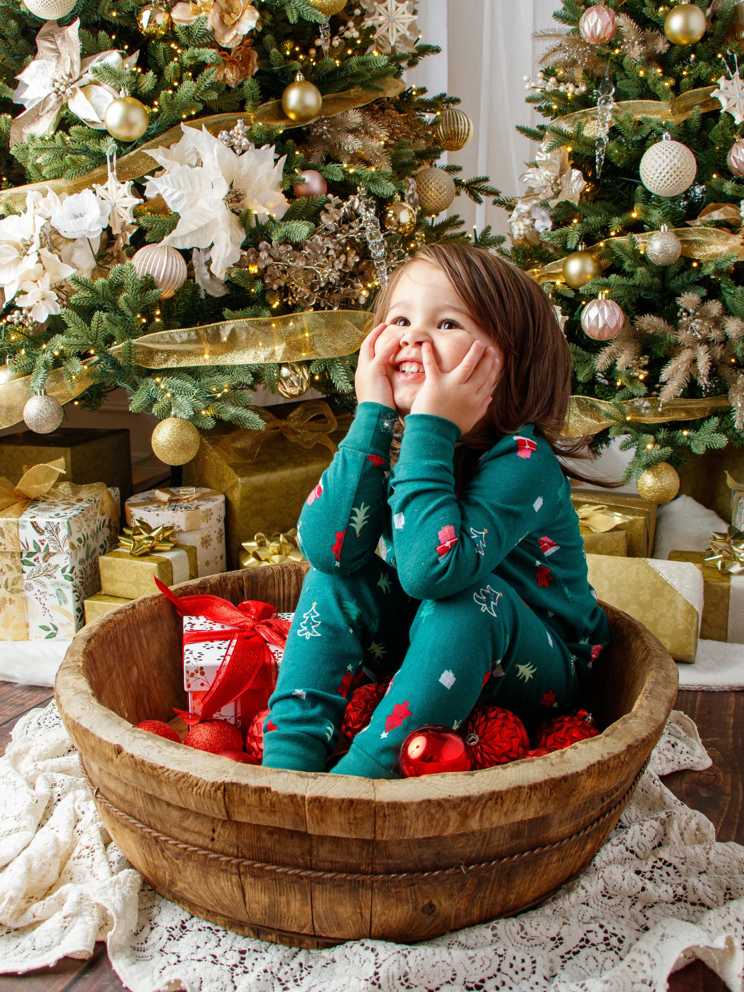 A young girl sitting in a wooden bowl filled with Christmas ornaments and gifts, smiling and holding her face with both hands, in front of decorated Christmas trees.  Milestone session, Redmond, WA.