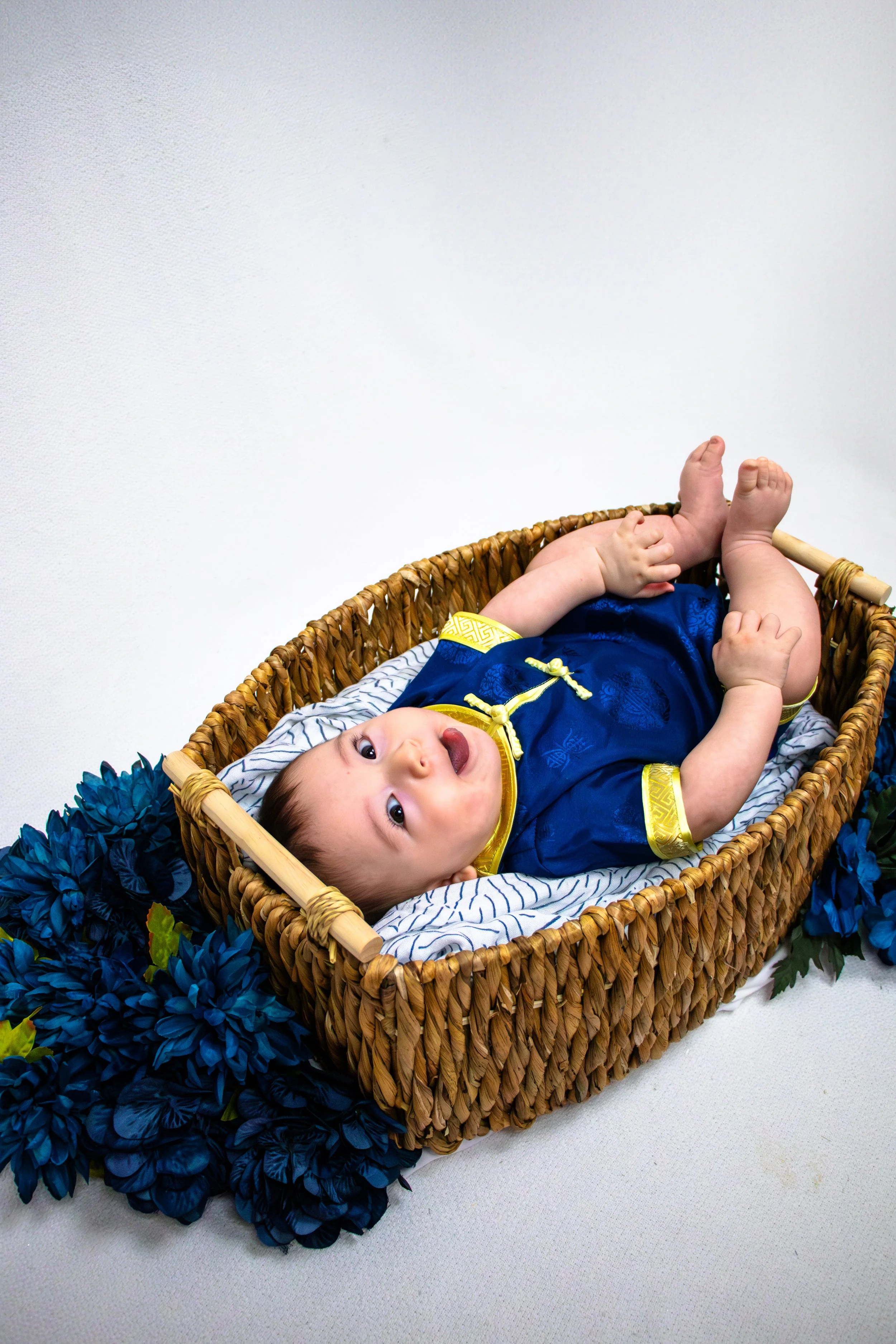 Baby lying in a woven basket, wearing a traditional blue and yellow outfit, surrounded by blue flowers. (Tummy Session)
