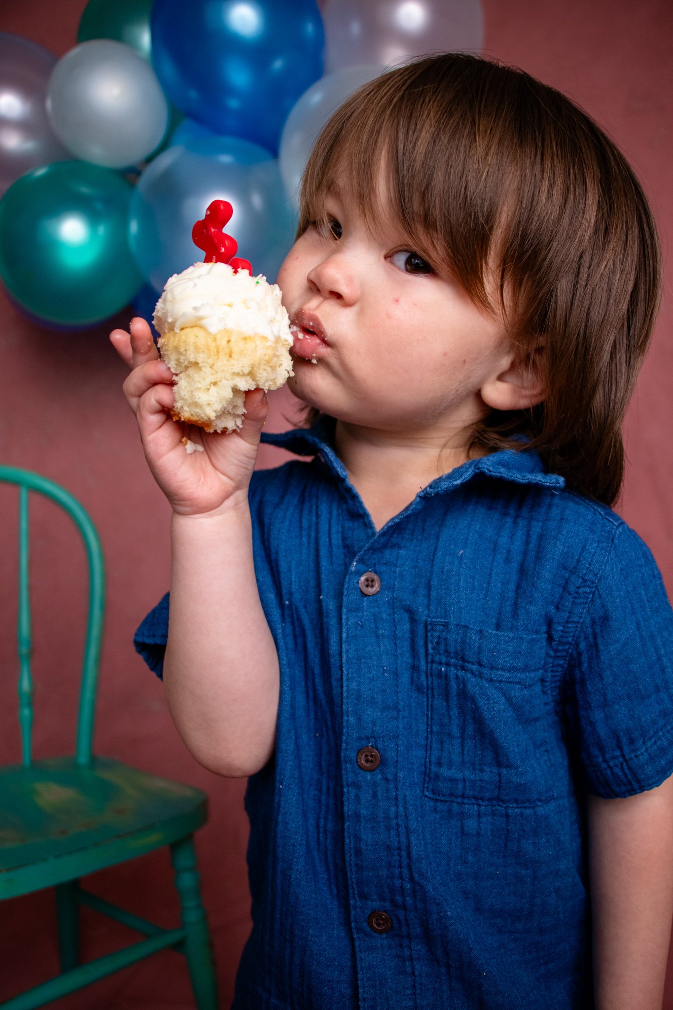 A young boy with brown hair in a blue shirt holding a cupcake with white frosting and a red number five candle, standing in front of a pink wall decorated with balloons.  Milestone session, Redmond, WA.