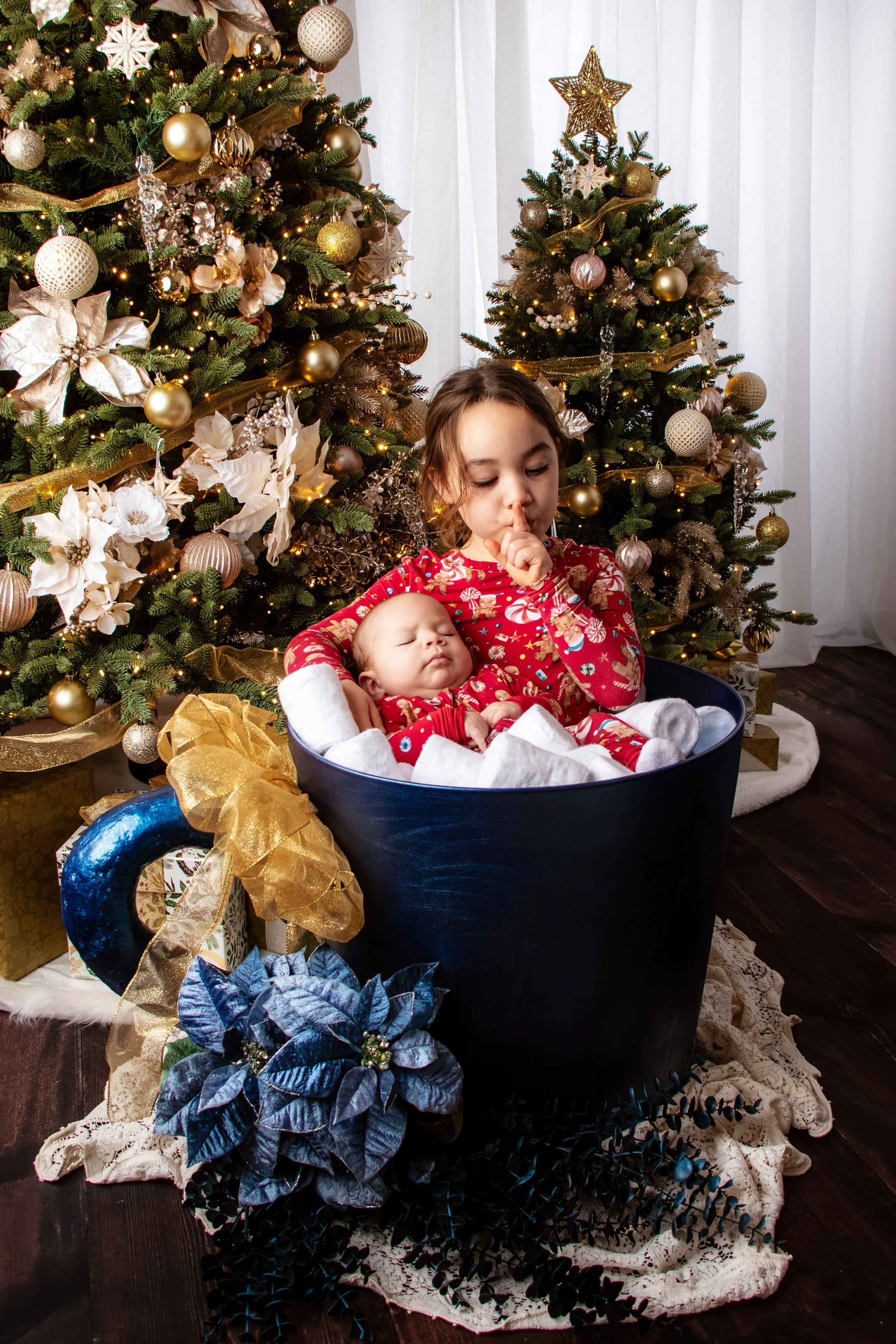 A young girl in festive pajamas sitting in a large navy blue bucket with a baby dressed in matching pajamas, in front of decorated Christmas trees with gold and white ornaments and lights.  Family session, Redmond, WA.