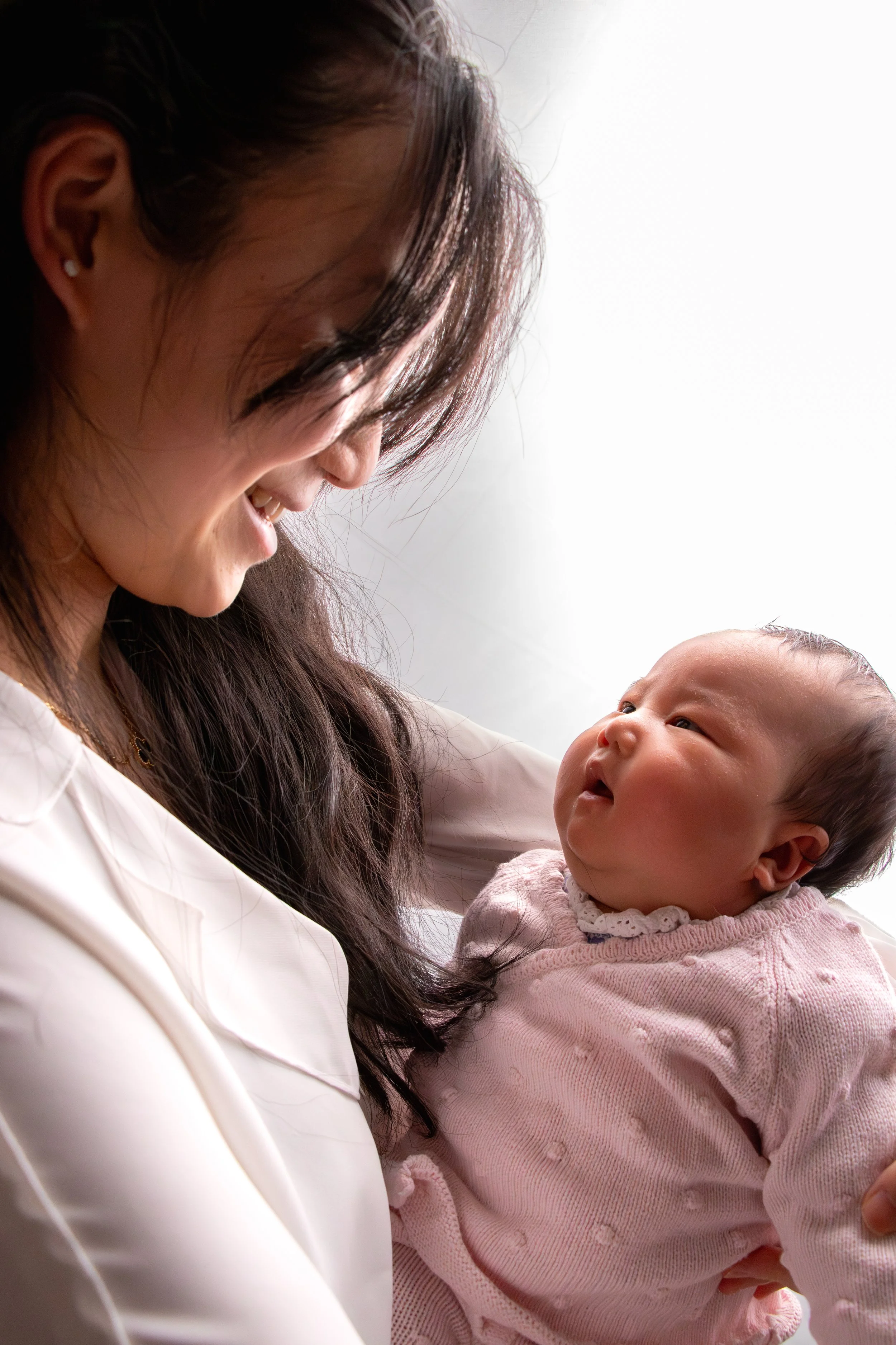 A woman smiling and looking at a baby girl she is holding, with both appearing happy and engaged. (Newborn Session)