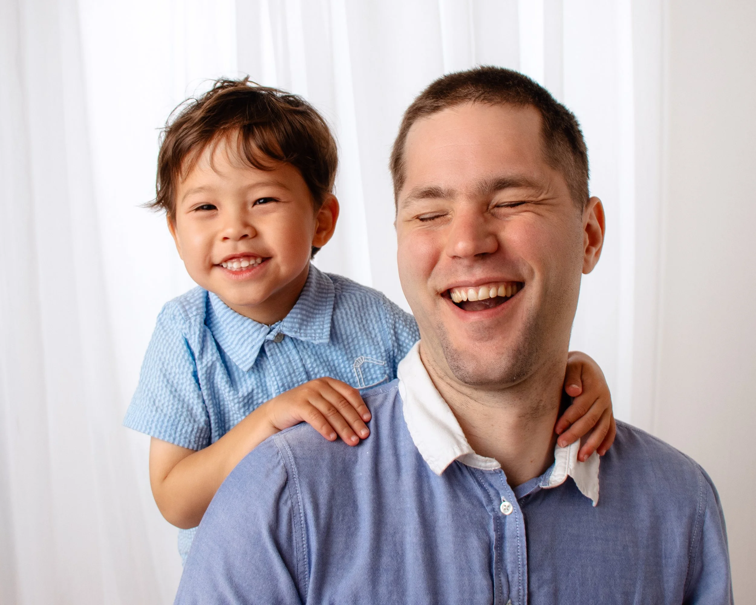 A young boy with brown hair smiling and a man with short brown hair laughing, with the boy's hand on the man's shoulder, both wearing blue shirts against a white background.  Family session, Redmond, WA.