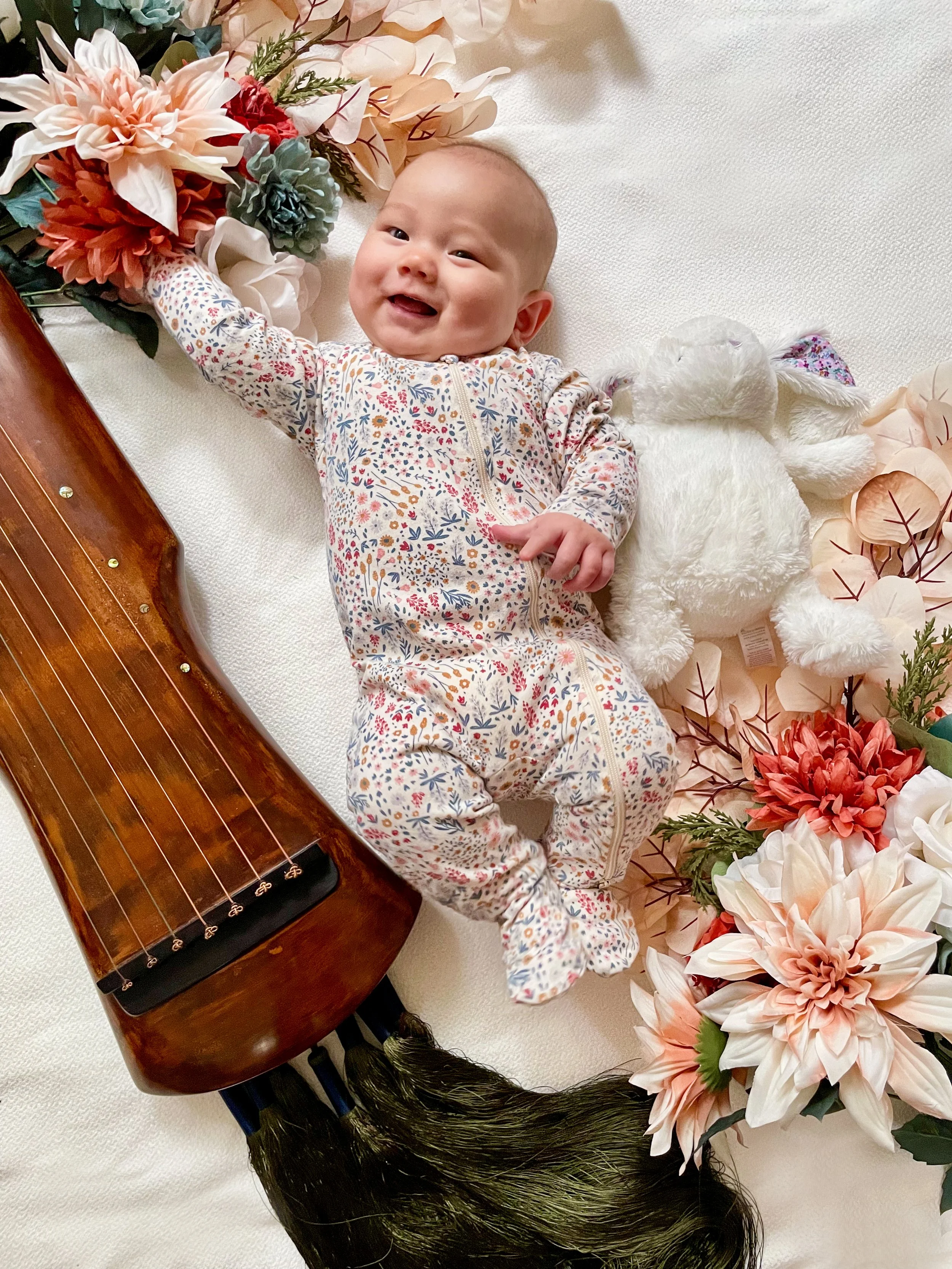Smiling baby lying on white blanket surrounded by flowers and plush toys, with a wooden stringed instrument and hair accessory nearby. (Mid-Autumn Festival Tummy Session)