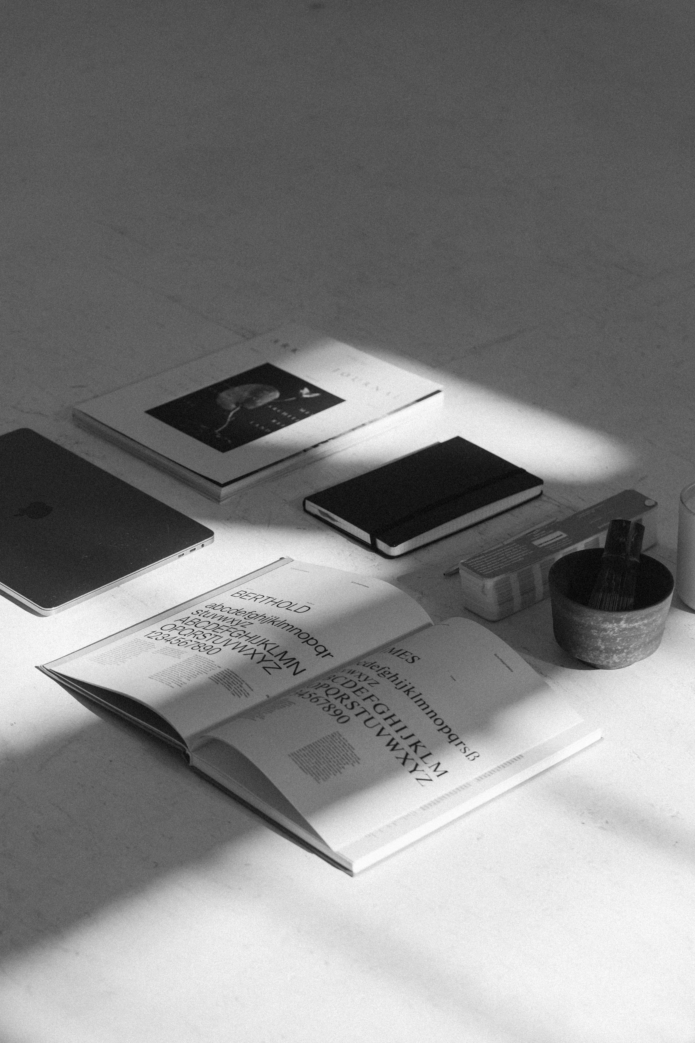 A woman sitting at a white table with a laptop, camera, and decorative items in a minimalistic room with white walls.