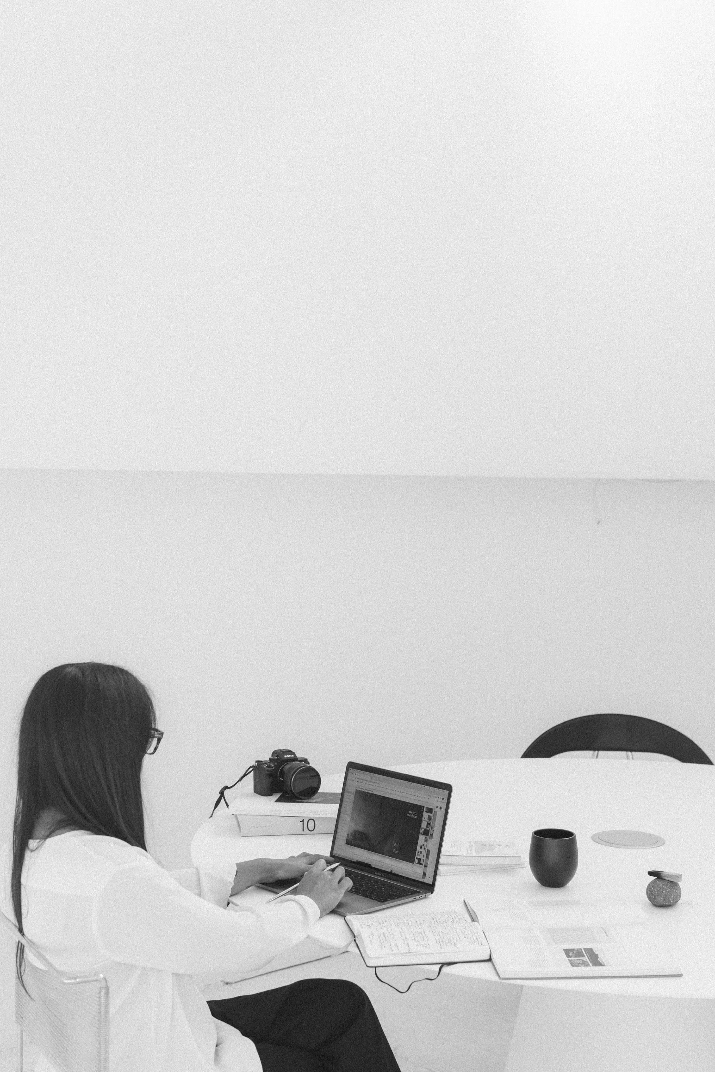 A woman sitting at a white table with a laptop, camera, and decorative items in a minimalistic room with white walls.