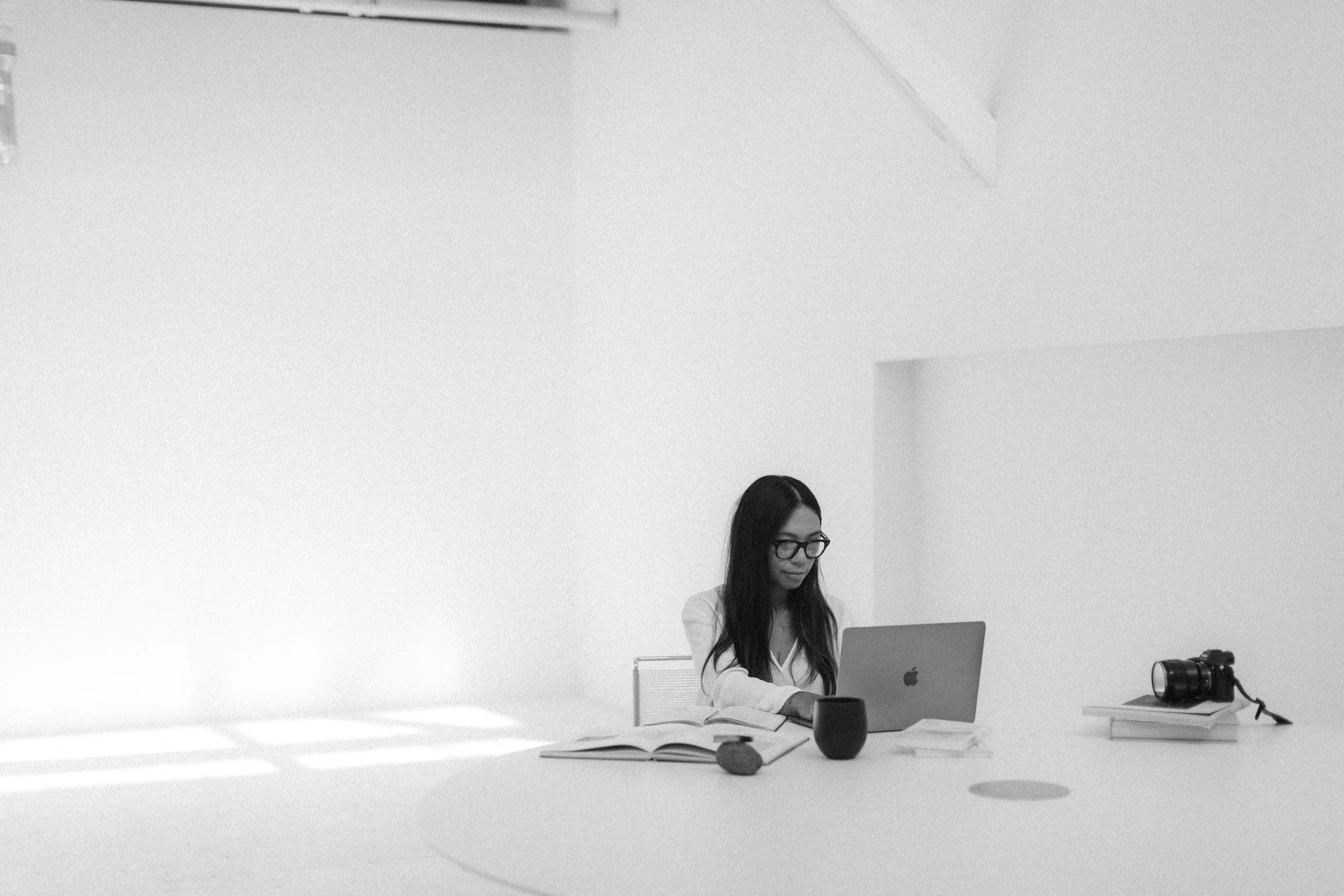 A white paper lantern lamp on a desk, with a small bowl, a magazine, a closed laptop, and glasses nearby, in black and white.