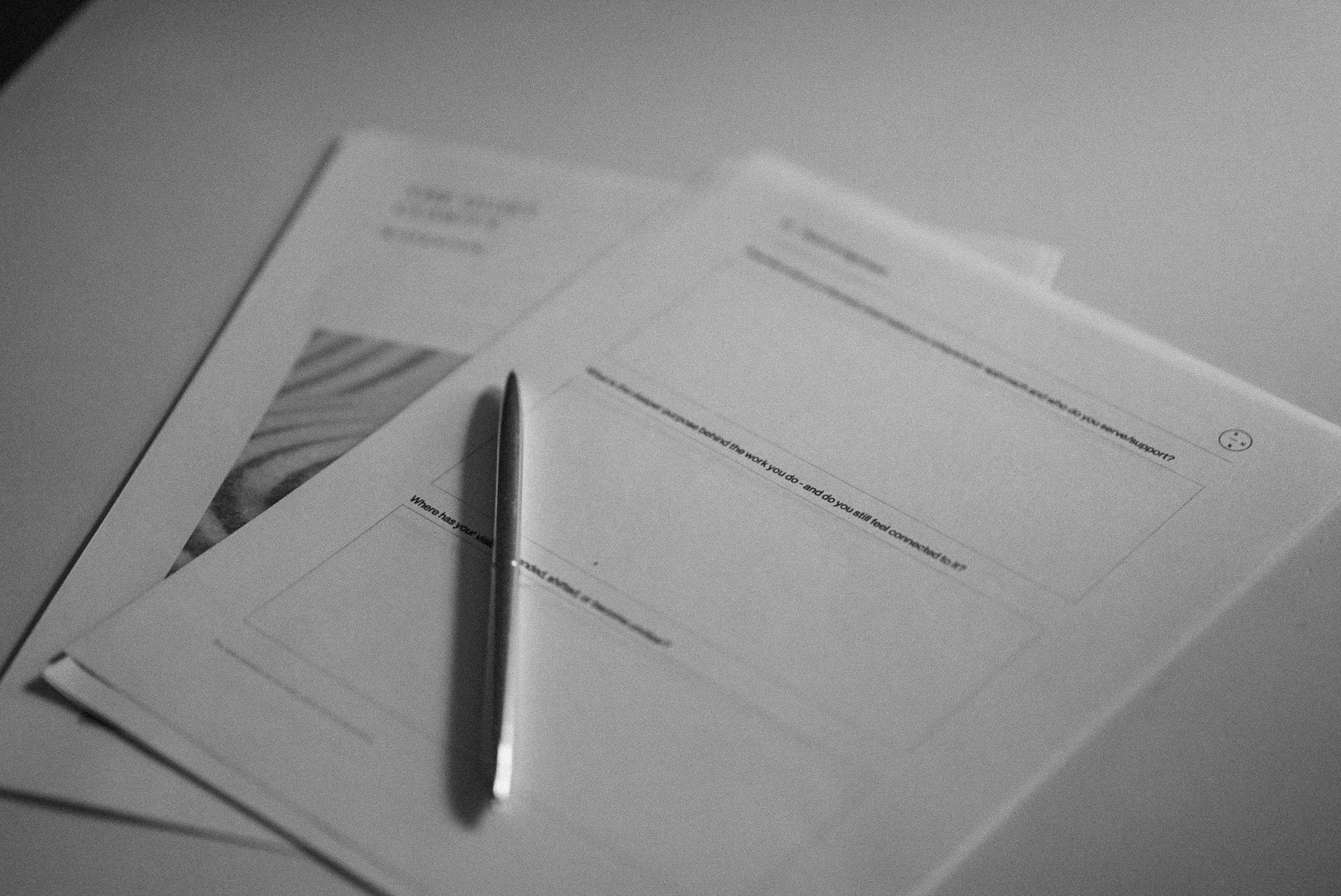 Black and white photograph of a desk with printed forms, a pen, and a tablet or laptop.