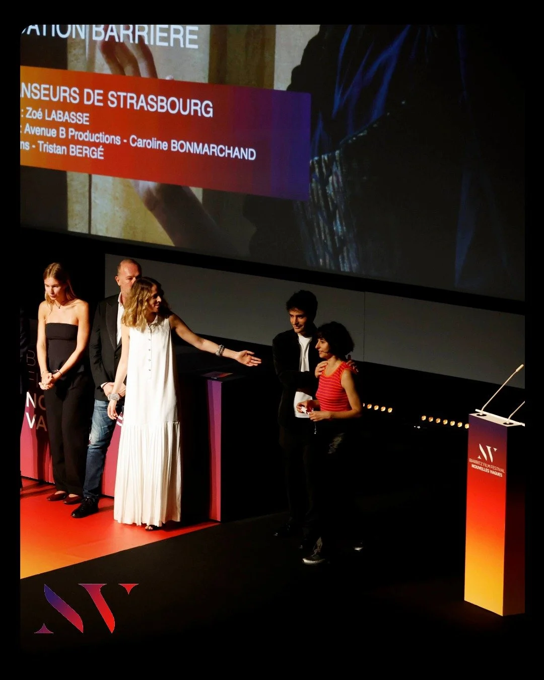 Scène d'une cérémonie de remise de prix lors d'un festival de cinéma, avec plusieurs personnes sur scène et un écran projetant un texte en français mentionnant les nominés. La scène est sombre avec un éclairage focalisé sur les intervenants.