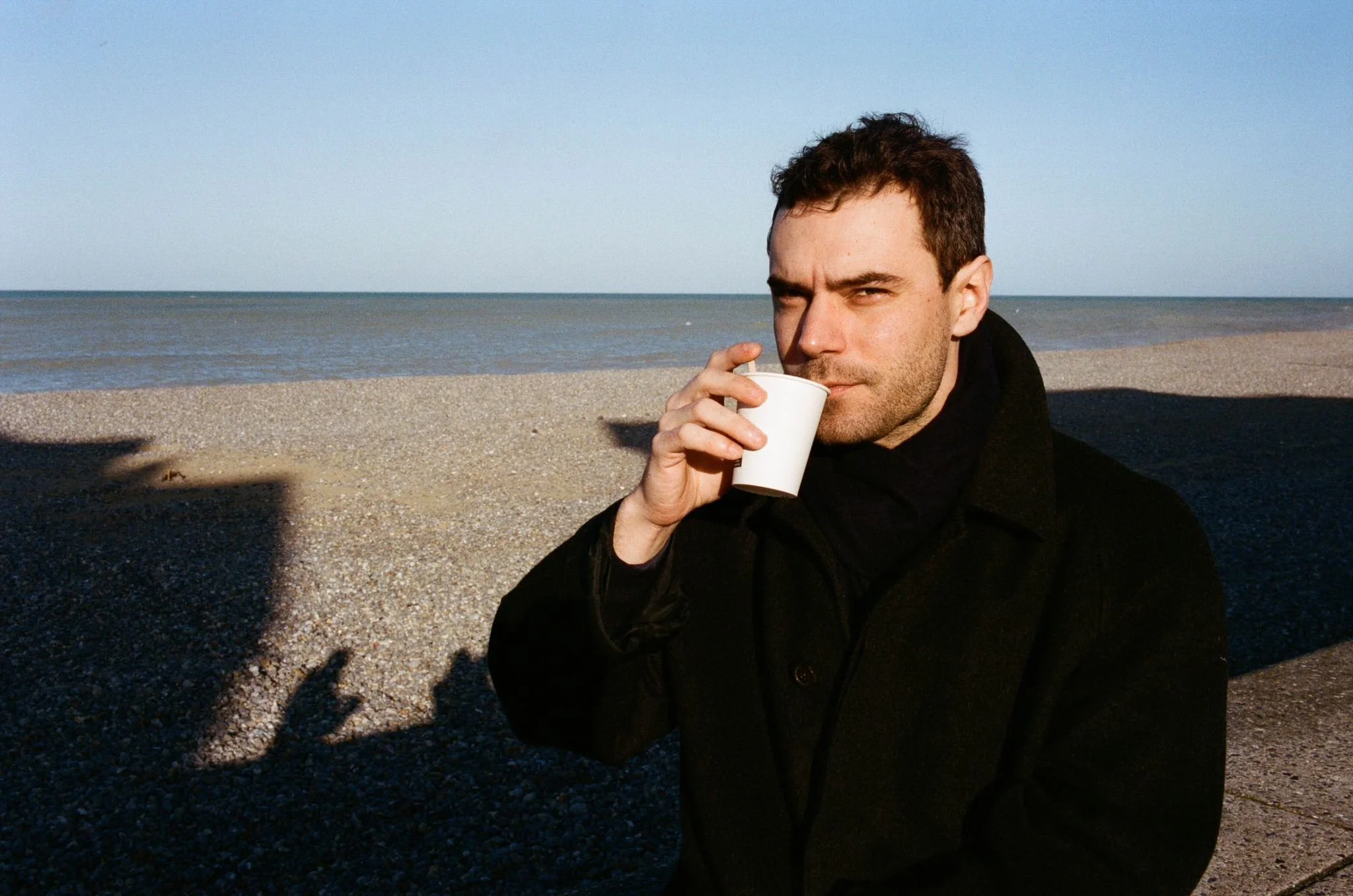 Un homme en manteau noir buvant dans un gobelet en carton sur une plage avec la mer en arrière-plan.