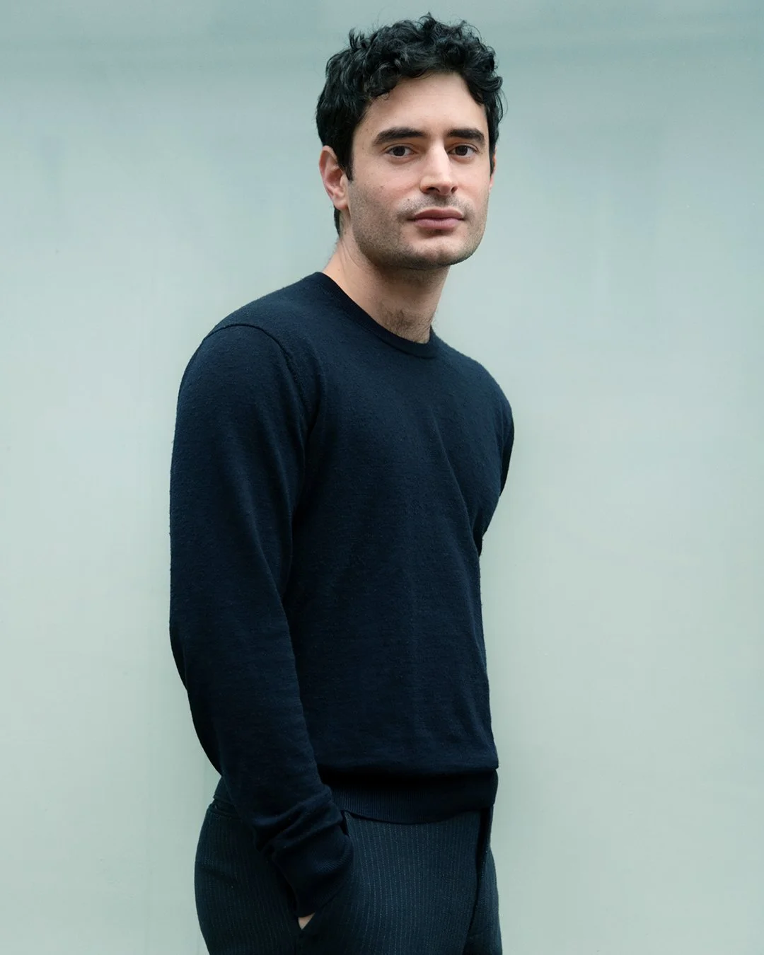 Portrait d'un homme avec des cheveux noirs, portant un pull noir, se tenant contre un fond gris clair.