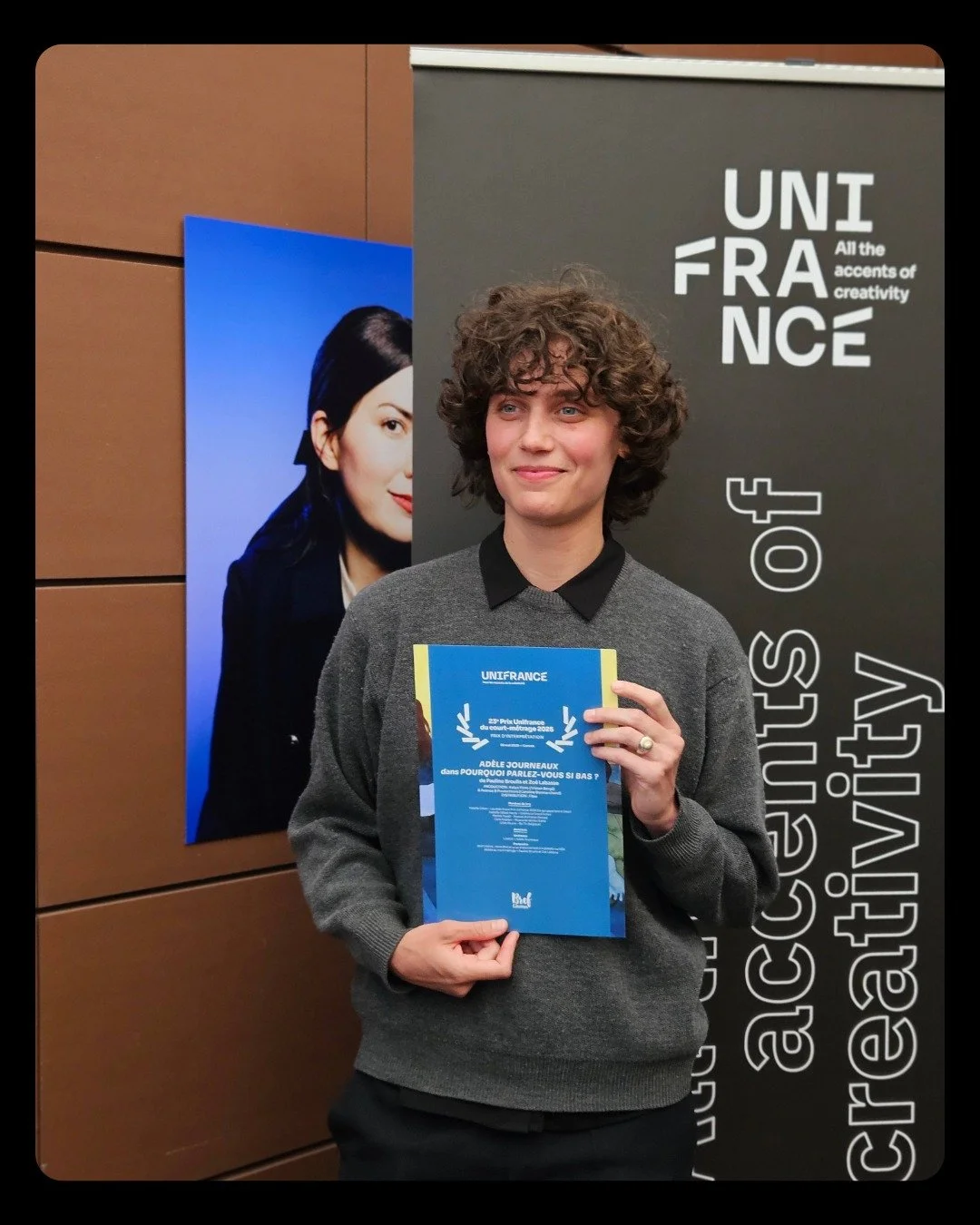 Femme souriante tenant un diplôme lors d'une cérémonie, avec un panneau noir à l'arrière-plan affichant 'UNIFRANCE' et des textes en anglais et français sur la créativité et l'éducation.