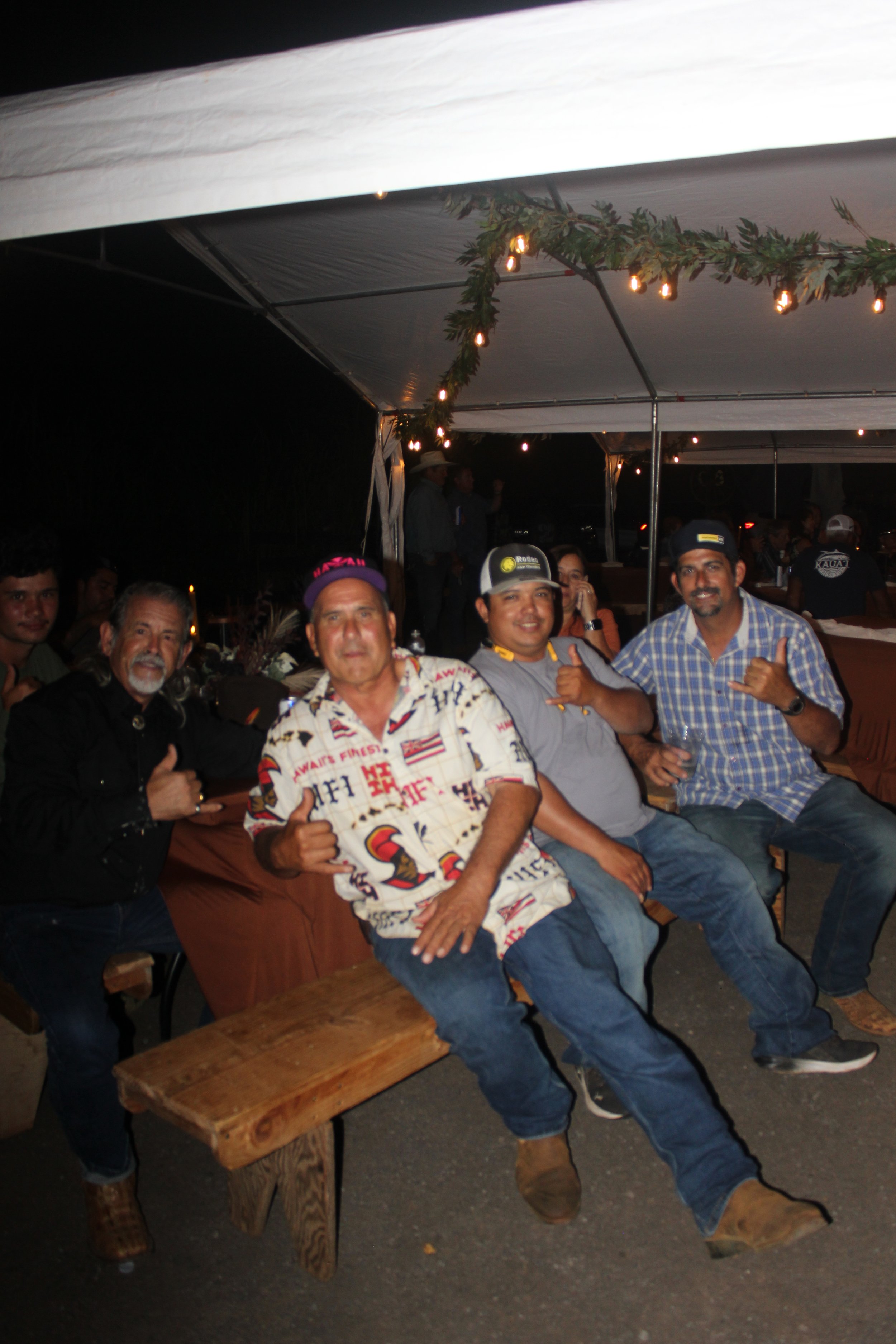 Group of men sitting on wooden benches under a decorated tent at night, smiling and making hand gestures.