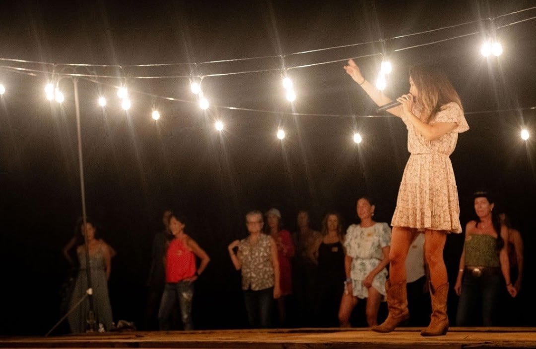 A woman in a pink floral dress and brown boots singing into a microphone on stage. Behind her, several people stand on a dark stage under string lights.