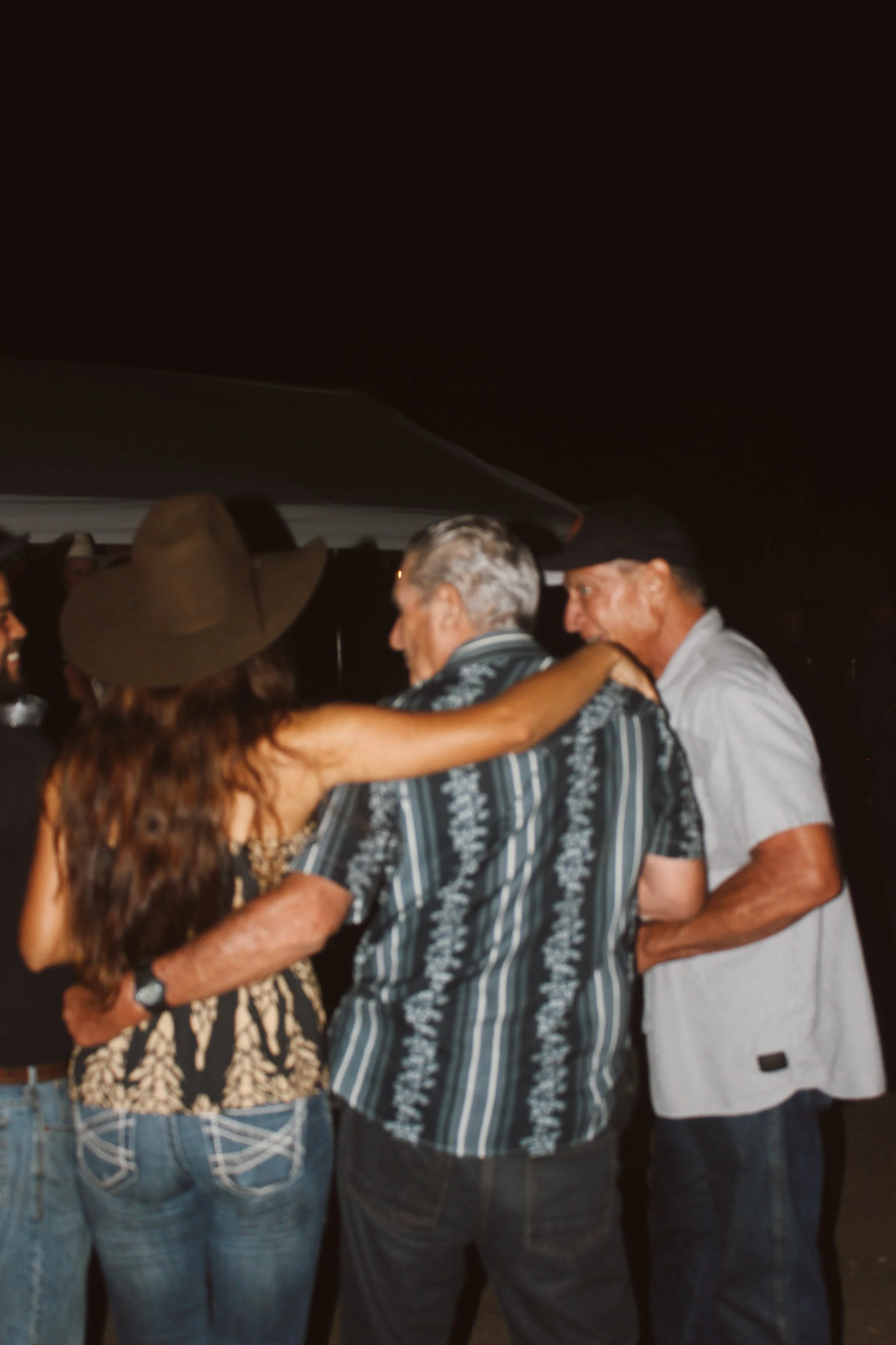 Group of people dancing and socializing at an outdoor evening event, including a woman with long hair and a hat, and three men, two of whom are embracing or holding each other.