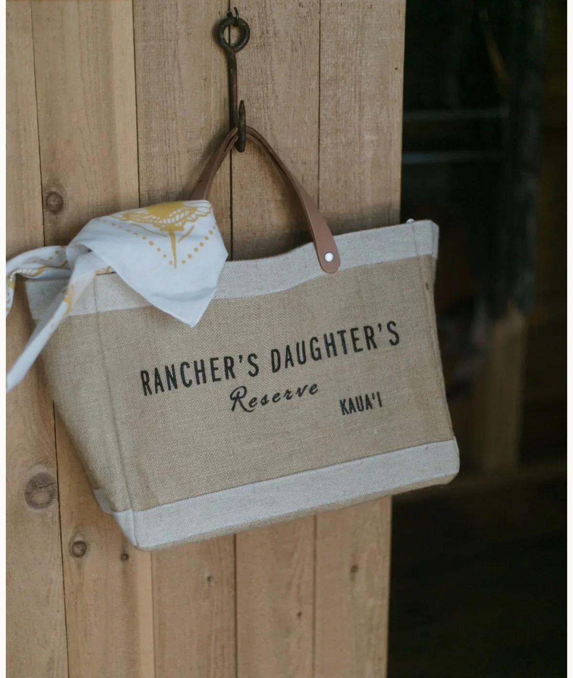 A beige canvas bag with leather handles hanging on a hook on a wooden wall, with the text 'Rancher's Daughter's Reserve Kauna' printed on it, and a white bandana with yellow design tied to one handle.