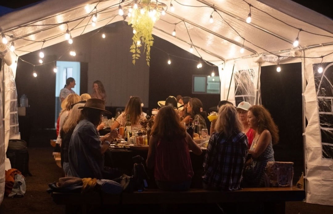 A group of people sitting at a long table under a tent decorated with string lights, enjoying a social gathering at night.