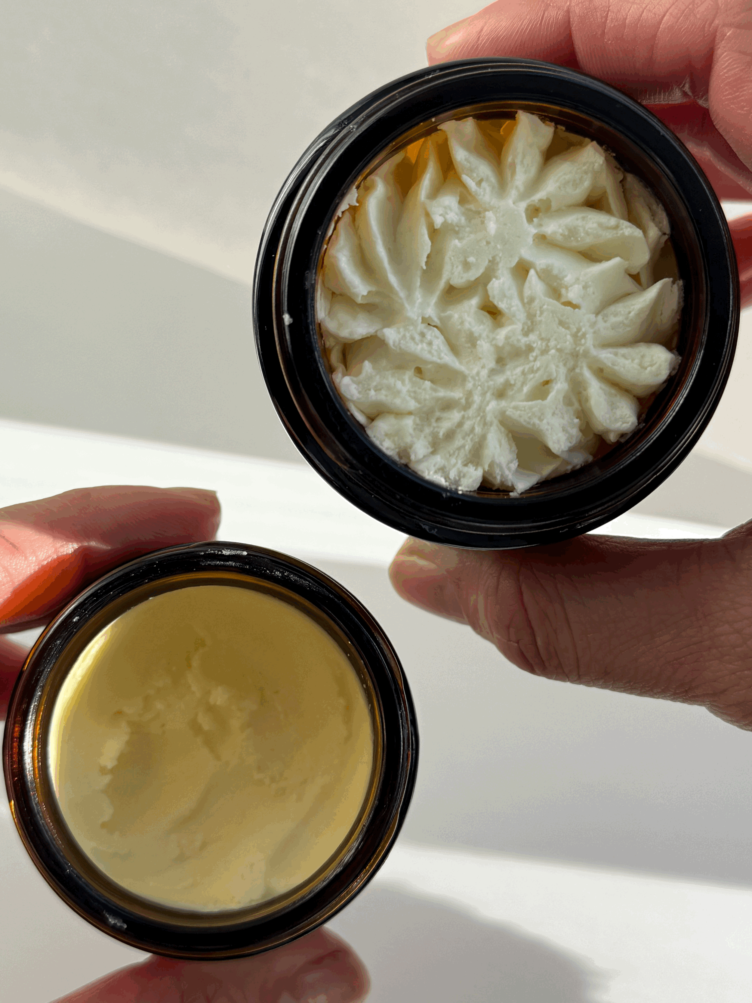 Two small jars held by hands, one containing a yellow wax or cream and the other filled with a cream or lotion topped with a decorative floral pattern.