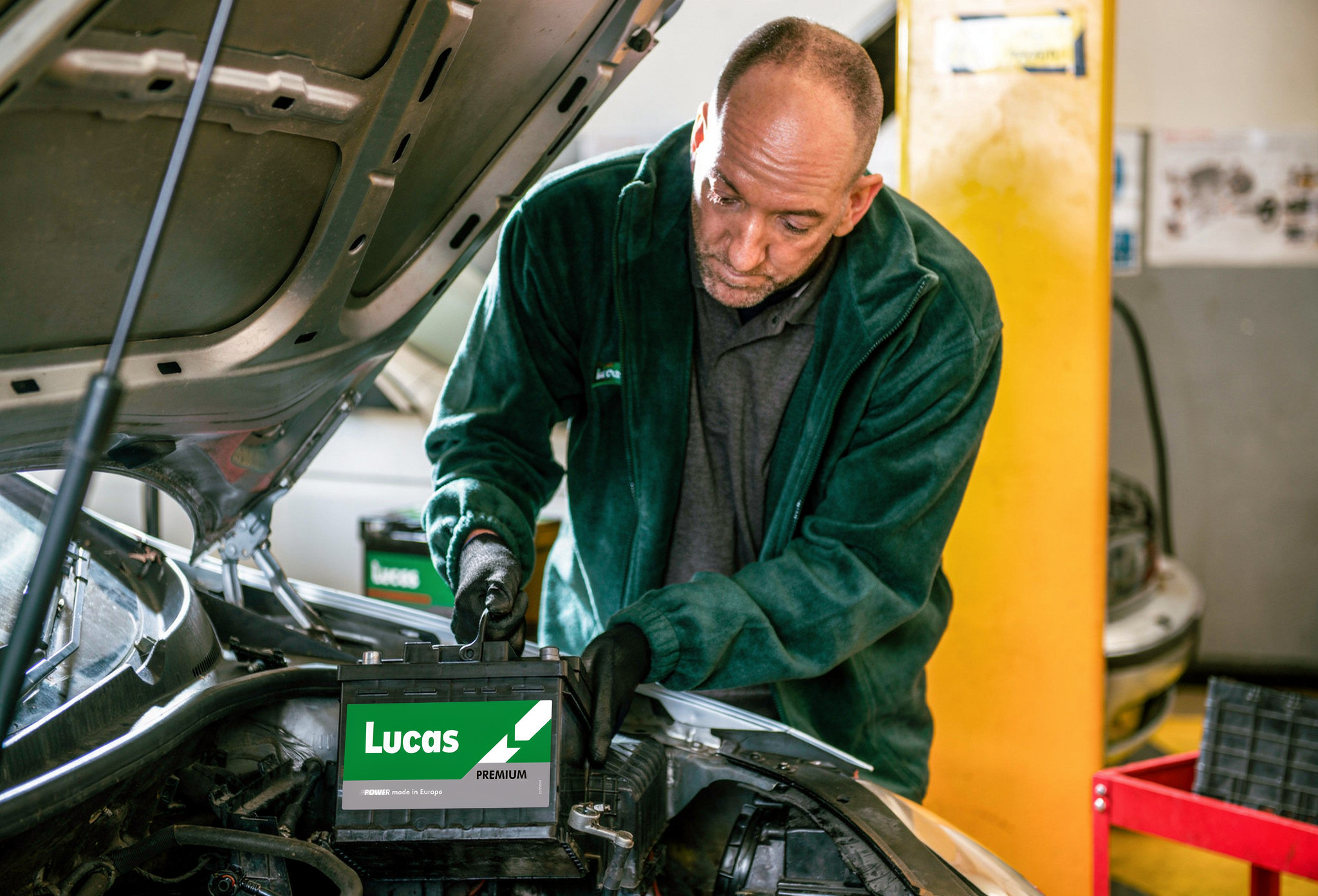 A mechanic in a green jacket works on a car battery under the open hood of a vehicle in a garage.