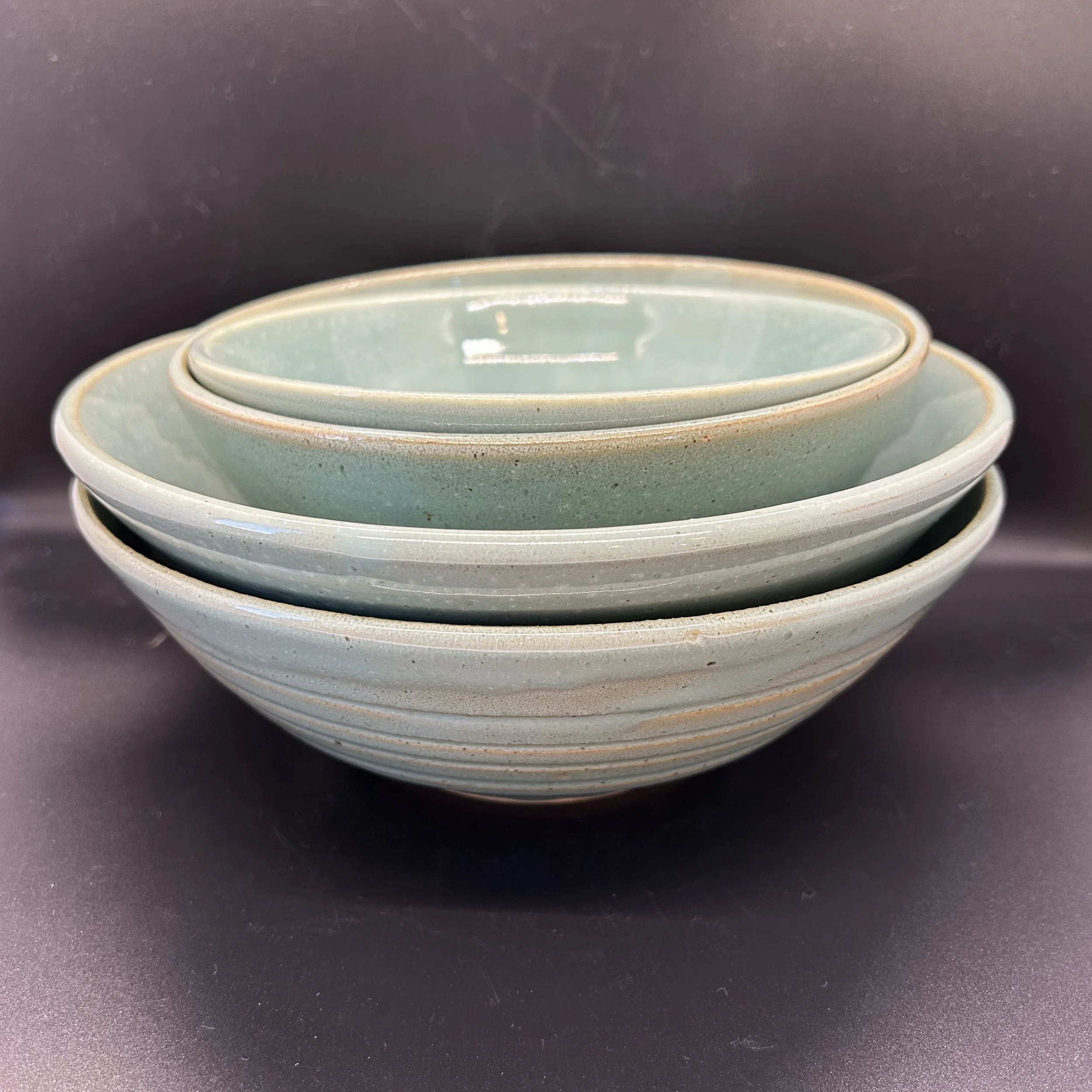 A set of four stacked ceramic bowls with a light greenish glaze on a dark surface.