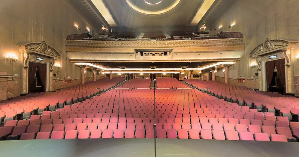 Empty theater auditorium with red seats, ornate gold and cream decor, and balconies on both sides.