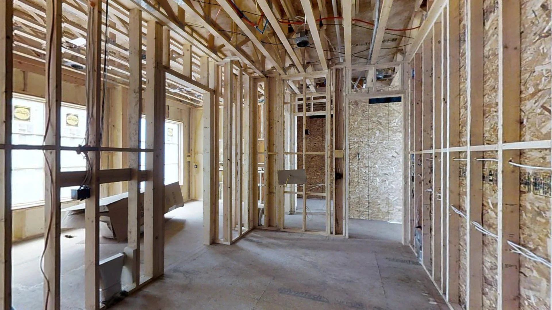 Interior of a house under construction showing wooden framing for walls and ceiling, with electrical wiring visible. Windows are installed, and the floor is unfinished.