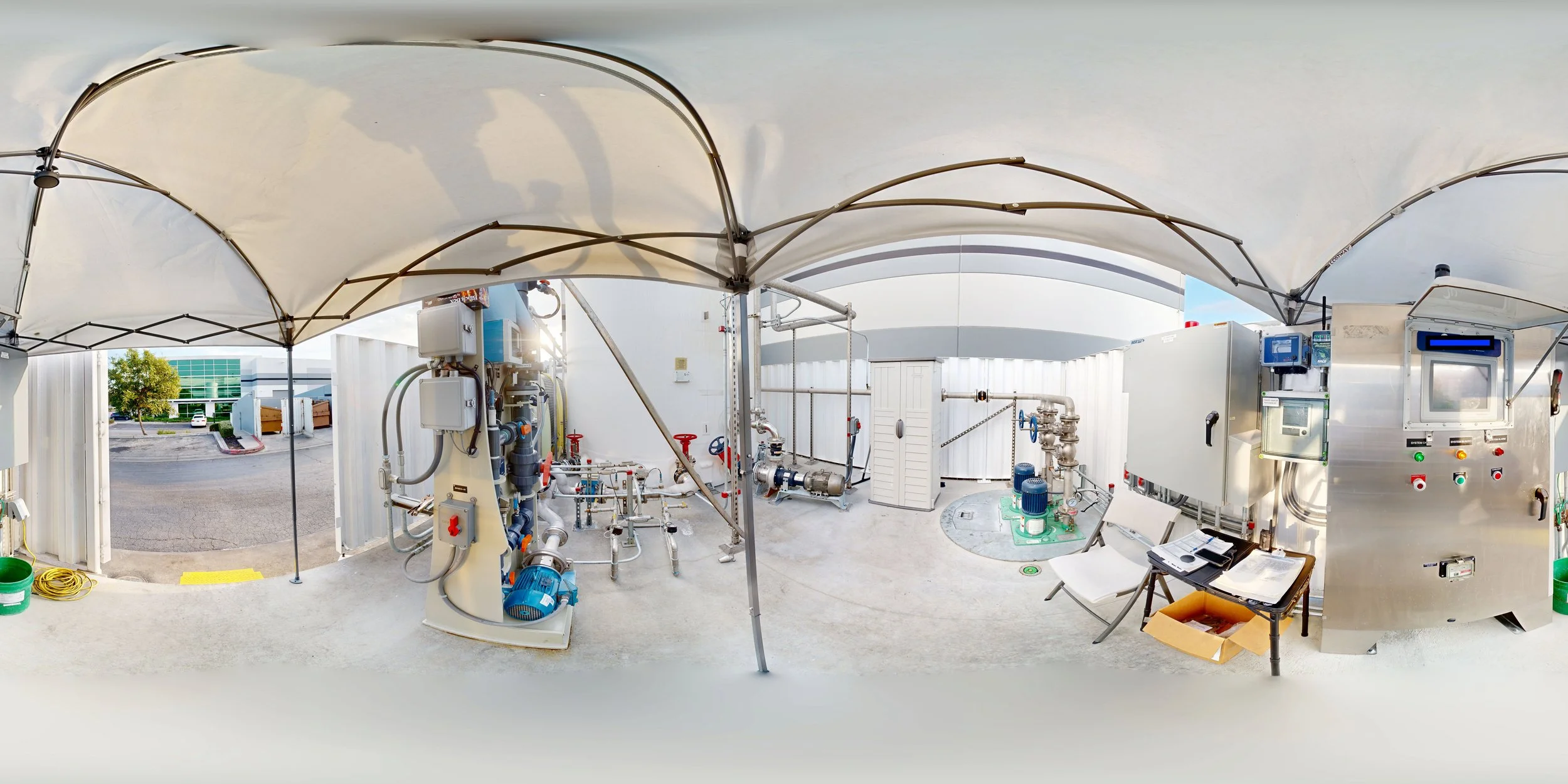 Wide-angle view of an outdoor industrial facility with pipes, valves, and control panels under a large canopy.