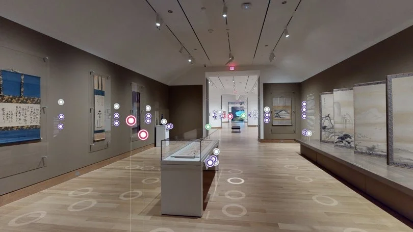 An art gallery exhibits paintings and scrolls on the walls, with a glass display case in the center. The room has wooden floors and a high ceiling with track lighting, leading to another section with more art displays.