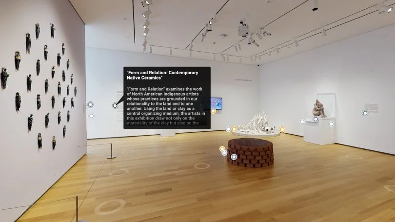 An art gallery with wooden floor and white walls displaying various sculptures and artworks. On the left wall, there are multiple small black sculptures arranged in a pattern. In the center, a brown circular stool is in front of a large black informational panel. To the right, there is a skeletal sculpture resembling a fish and a carved stone sculpture of a seated figure on pedestals. The ceiling has track lighting illuminating the exhibits.
