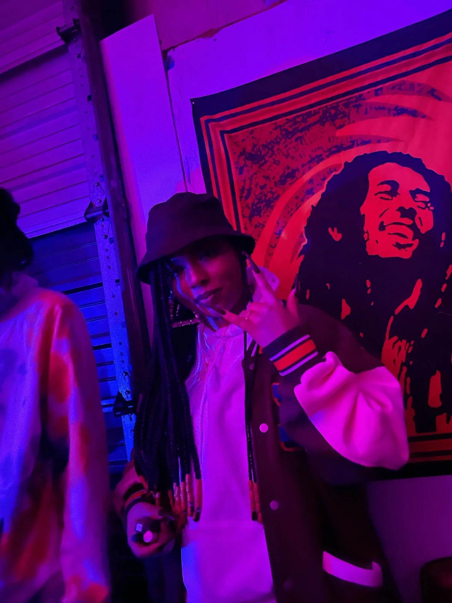 A young person with long braids wearing a bucket hat, a varsity jacket, and holding a phone, making a gesture with their hand in front of a wall with a colorful poster of Bob Marley in a neon-lit setting.