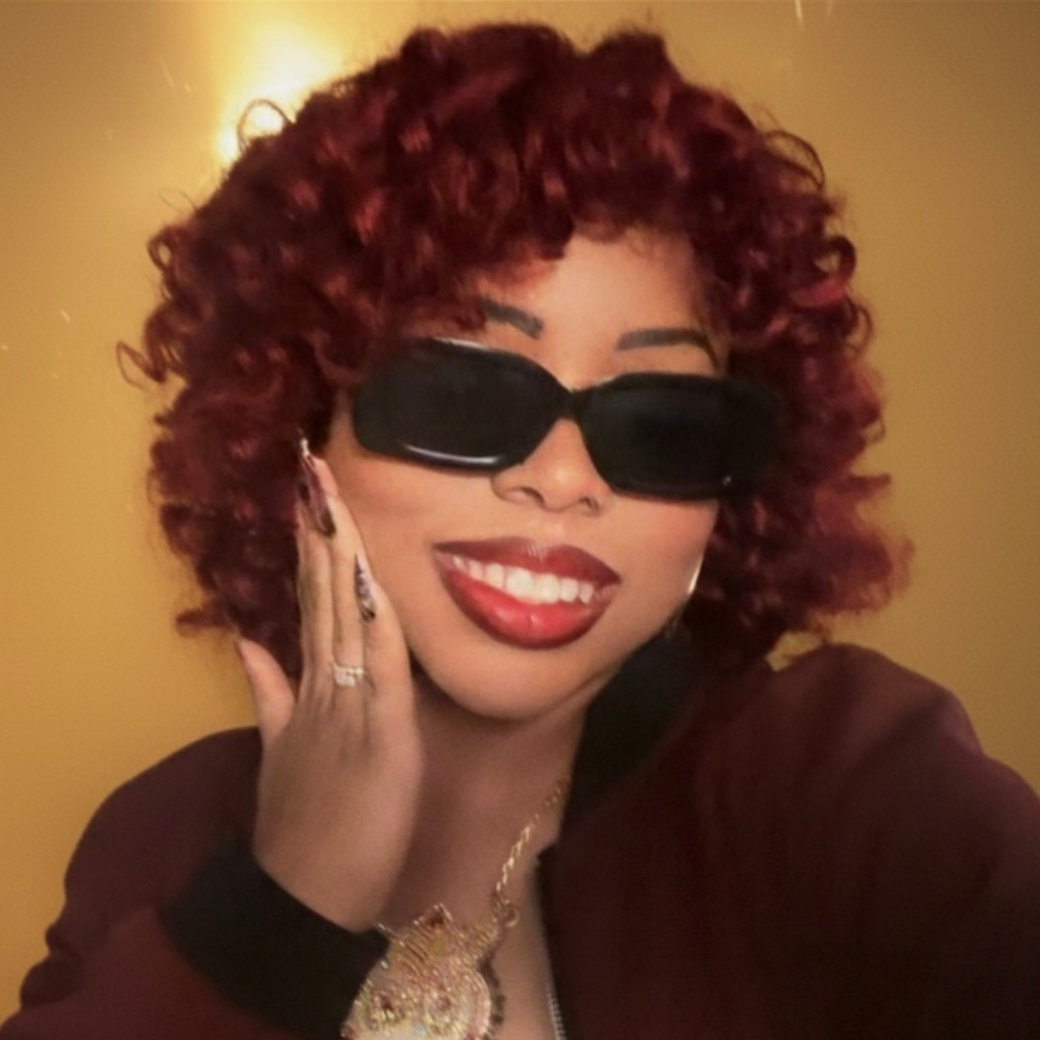 Mani Da Brat independent female hip-hop and R&B artist with curly red hair wearing sunglasses and burgundy jacket against yellow background