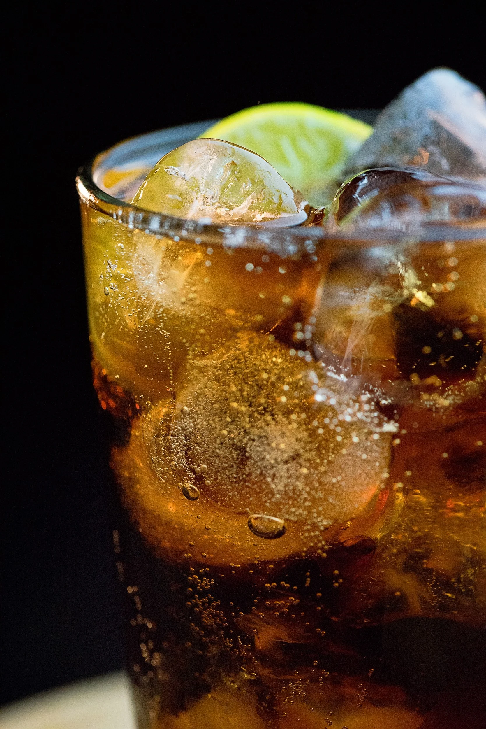 Close-up of a glass of cola with ice and lemon wedge, with bubbles and condensation, against a black background.